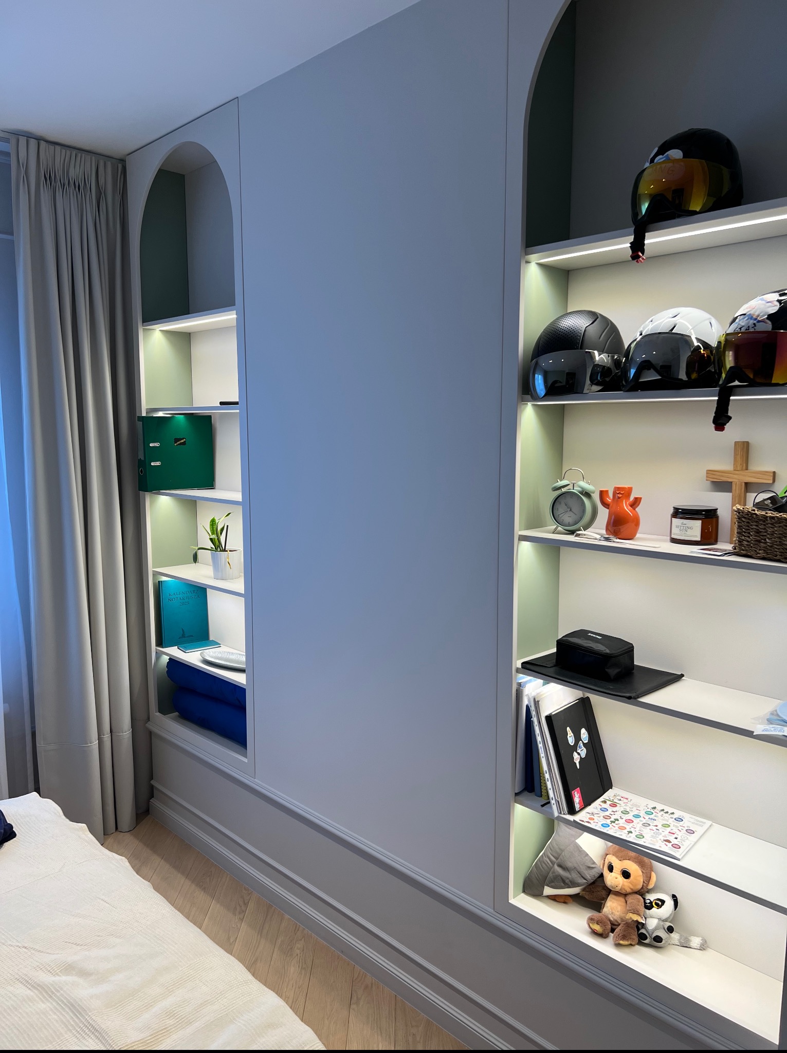 Biała szafa wnękowa z półkami podświetlonymi LED, wnęki w kształcie łuków, na półkach dekoracje, książki i kaski motocyklowe. Widoczny fragment łóżka.