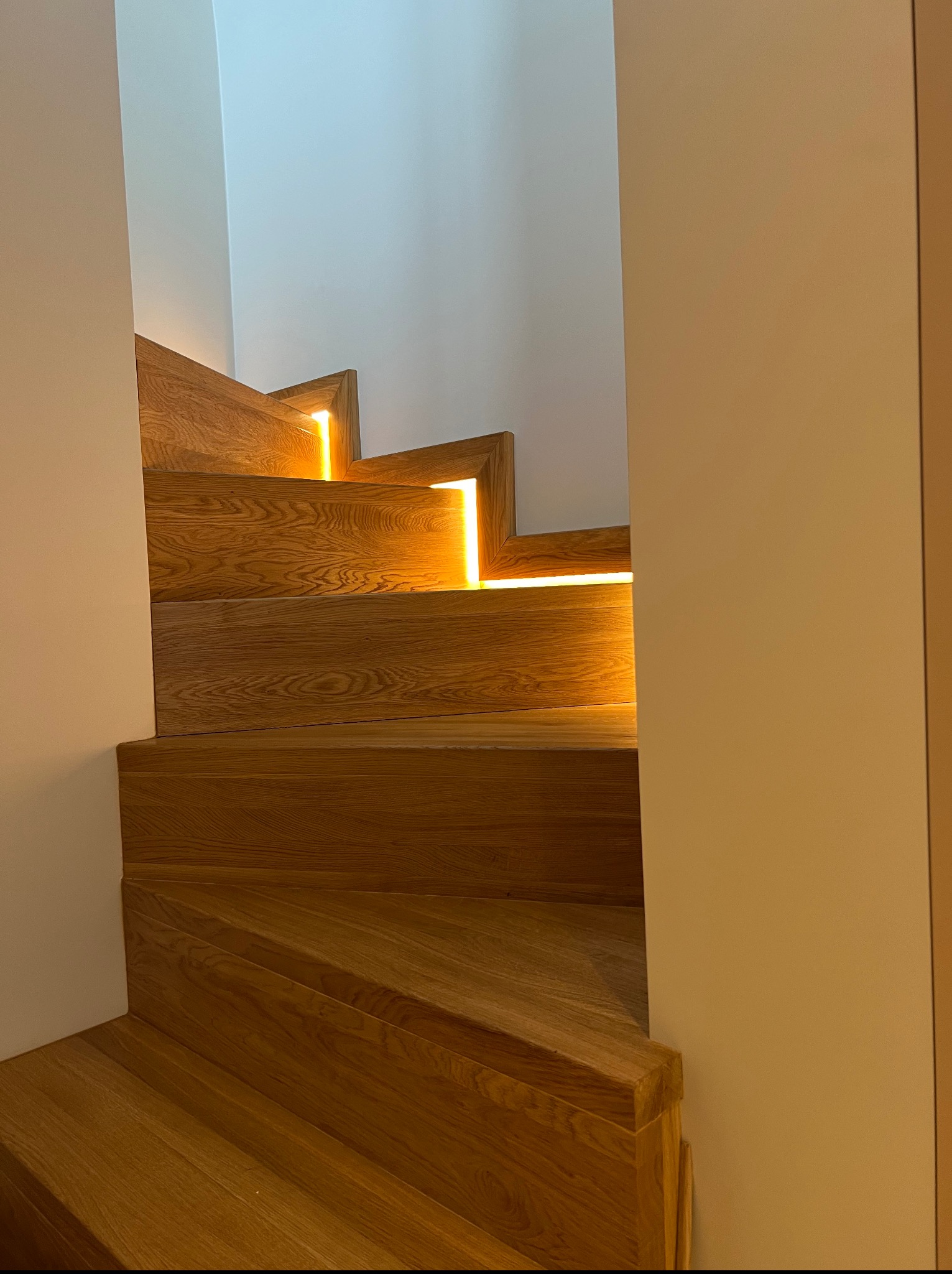 Drewniane schody z podświetleniem LED między stopniami, widok z dołu, oświetlenie ciepłe, minimalistyczne wnętrze z białymi ścianami, nowoczesny design.