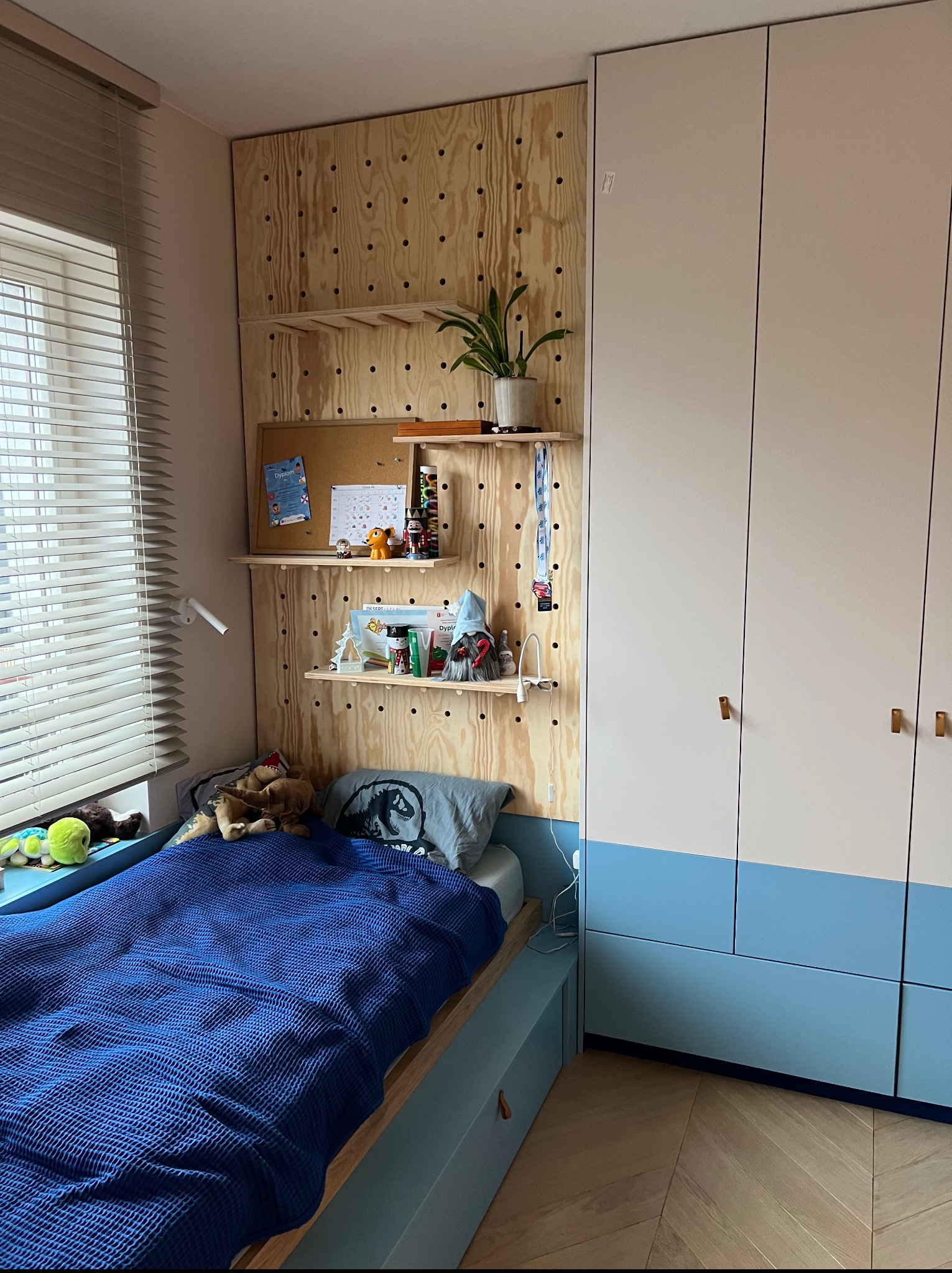 Pokój dziecięcy z szafą wnękową w kolorach bieli i błękitu, drewnianą ścianą z półkami i łóżkiem. Na półkach książki, zabawki i roślina doniczkowa. Jasne wnętrze.