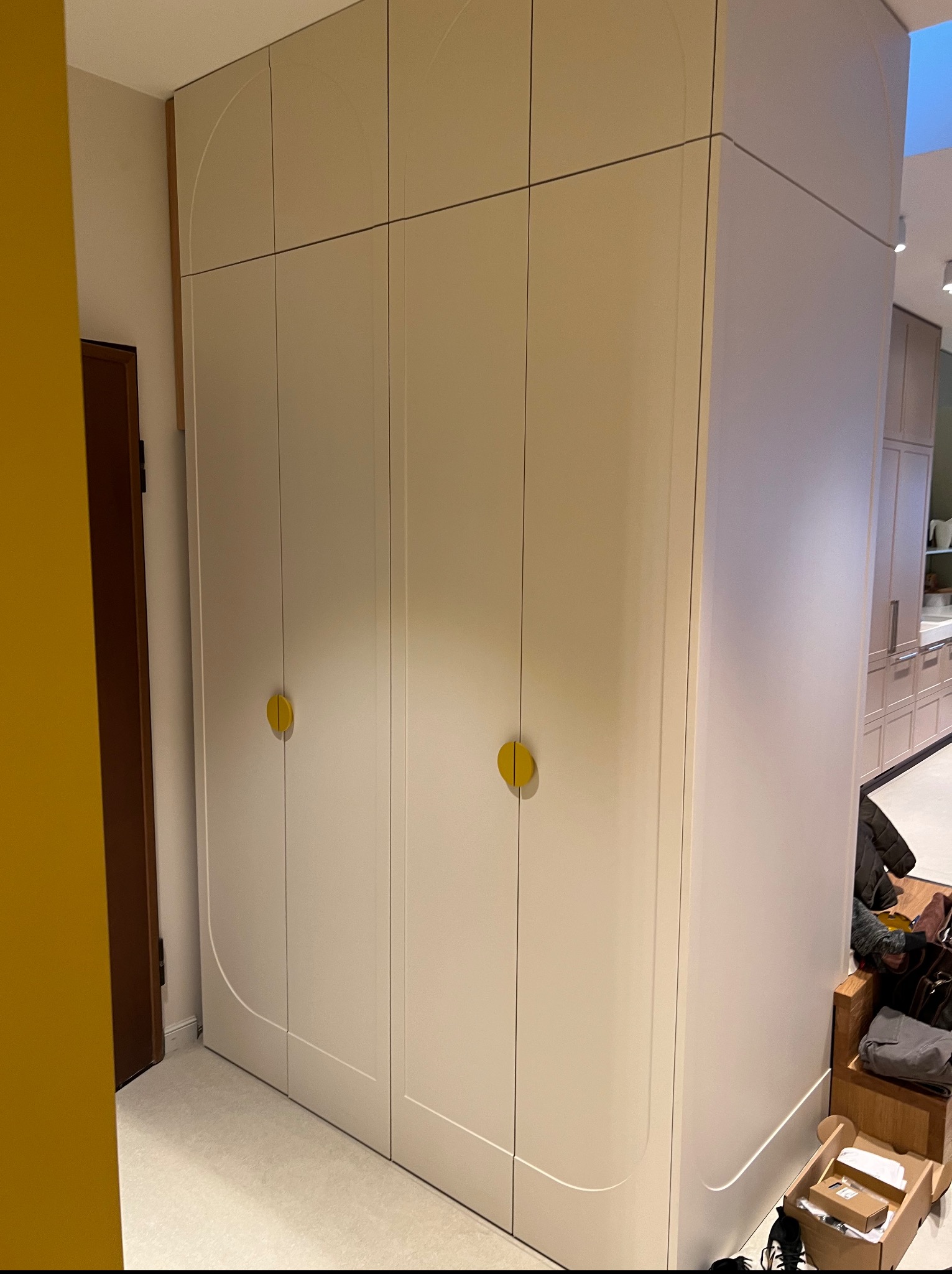 Biała szafa wnękowa z zaokrąglonymi krawędziami i żółtymi okrągłymi uchwytami, widoczna na tle jasnego wnętrza z elementami zabudowy kuchennej.