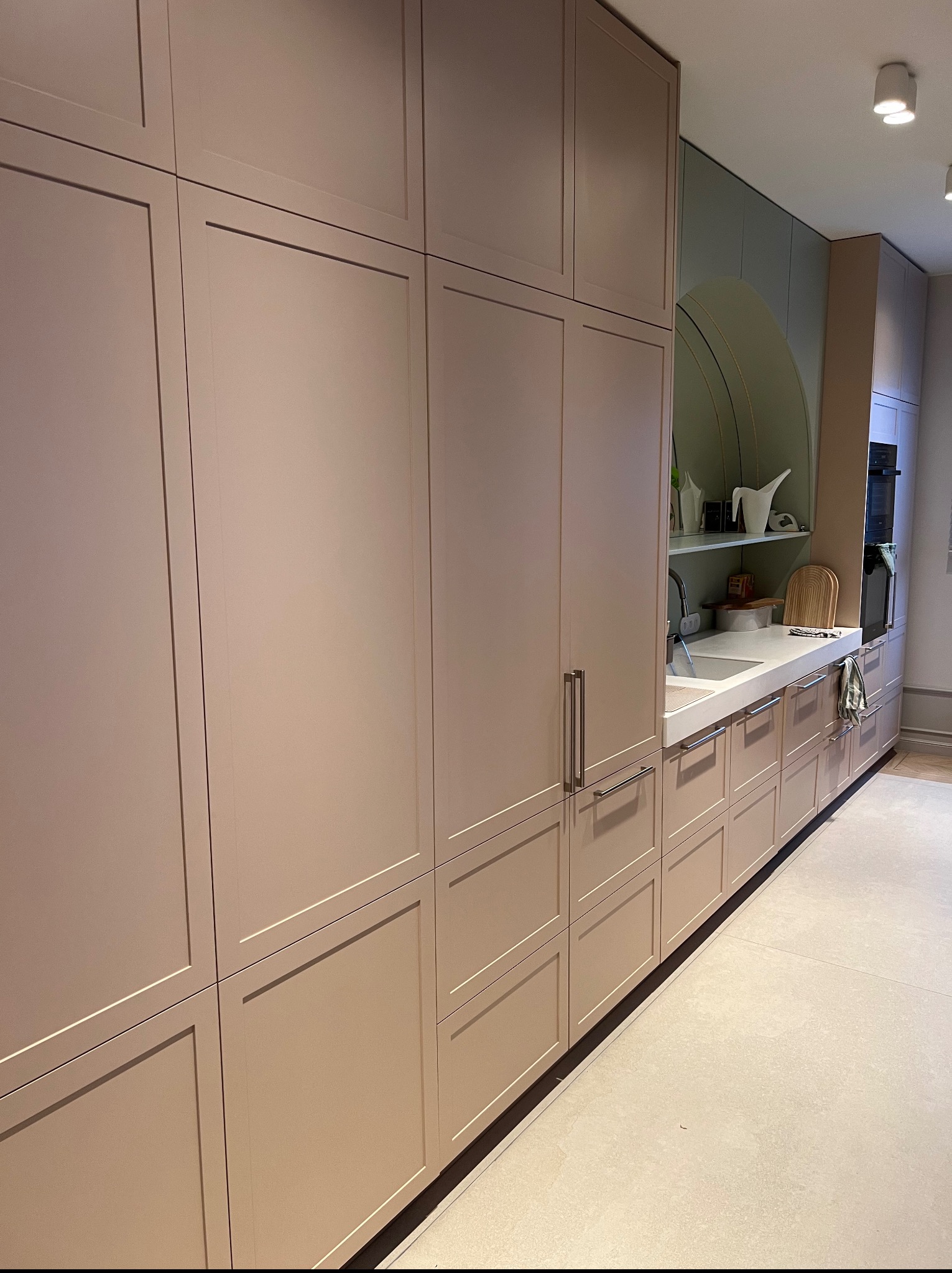 Jasna kuchnia z zabudową na wymiar. Fronty szafek w odcieniach beżu, minimalistyczne uchwyty. Na blacie akcesoria kuchenne i zlew. Nowoczesny design.