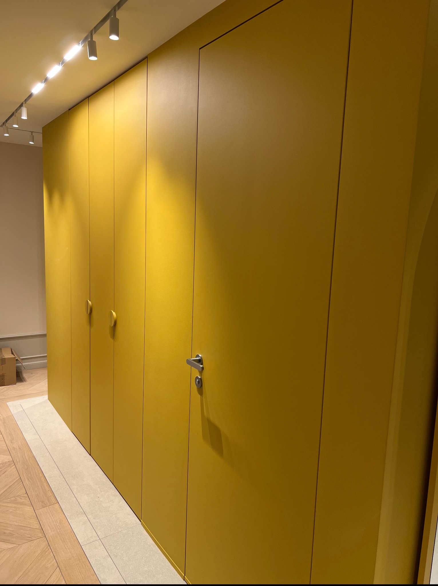 Żółta szafa wnękowa z dyskretnymi uchwytami i drzwiami bez widocznych zawiasów, wkomponowana w nowoczesne wnętrze z oświetleniem punktowym.