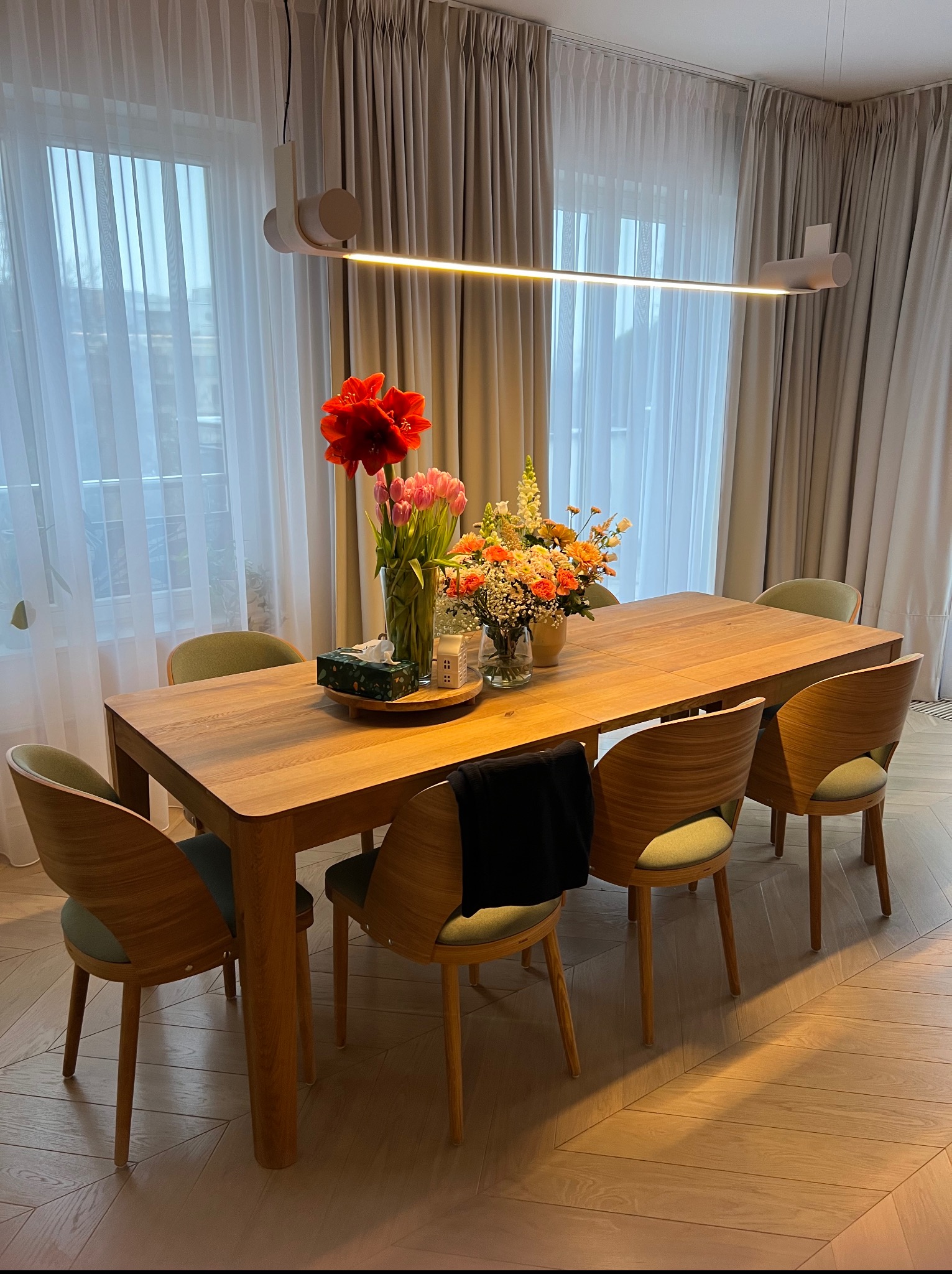 Drewniany stół z krzesłami w jadalni, bukiety kwiatów w wazonach, minimalistyczna lampa. Parkiet ułożony we wzór jodełki. Stonowana kolorystyka, naturalne światło.