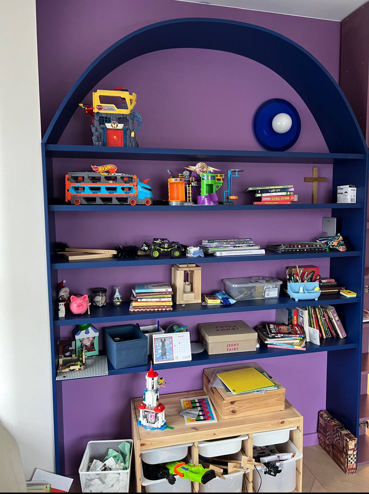 Niebieski regał z półkami w pokoju dziecięcym, wypełniony zabawkami, książkami i dekoracjami na tle fioletowej ściany. Półki w kształcie łuku.
