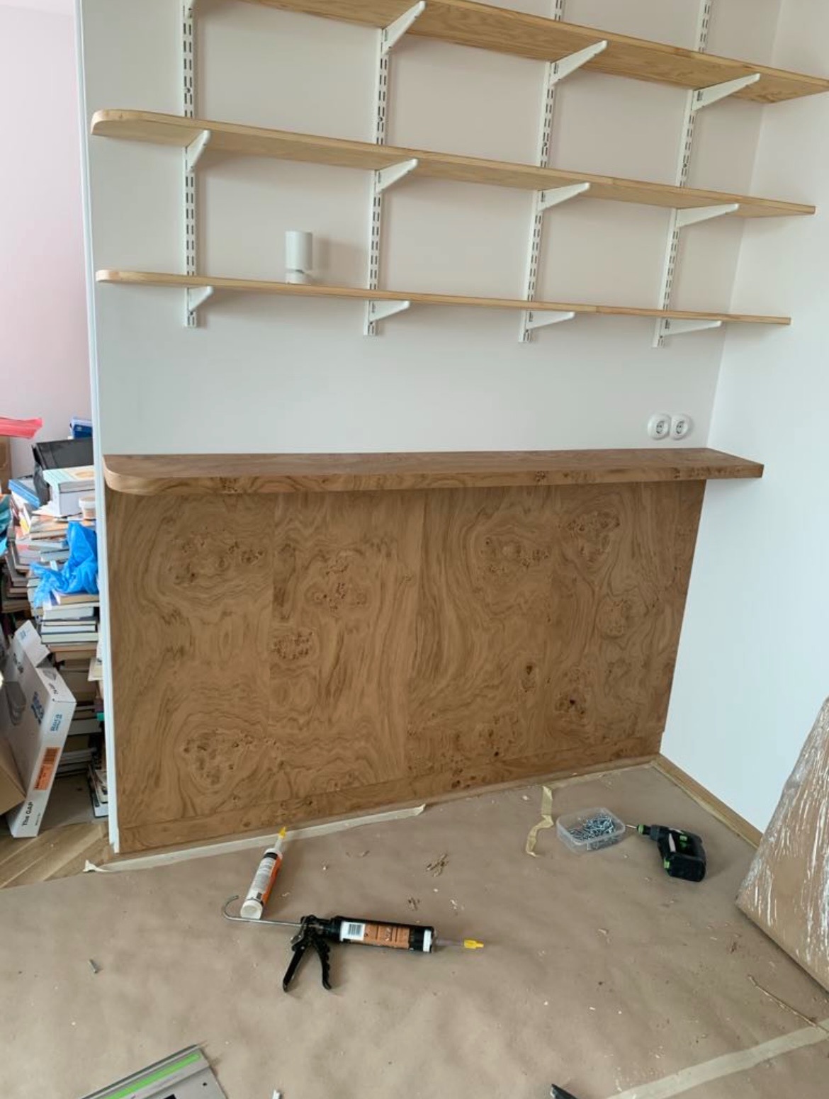 Montaż drewnianej zabudowy pod półkami na ścianie. Widoczne narzędzia i materiały montażowe. Wykończenie z forniru o ciekawym wzorze.