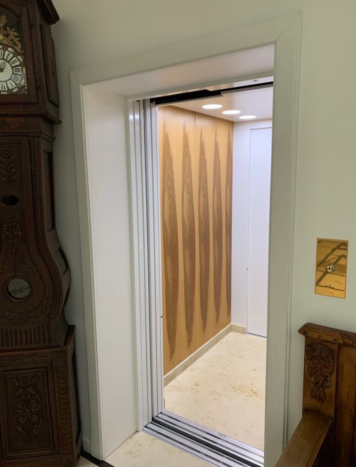 Wnętrze windy z drewnianą okładziną ścian, widoczne przez białą ramę drzwi. Obok stoi stary zegar i krzesło. Jasne oświetlenie wewnątrz.