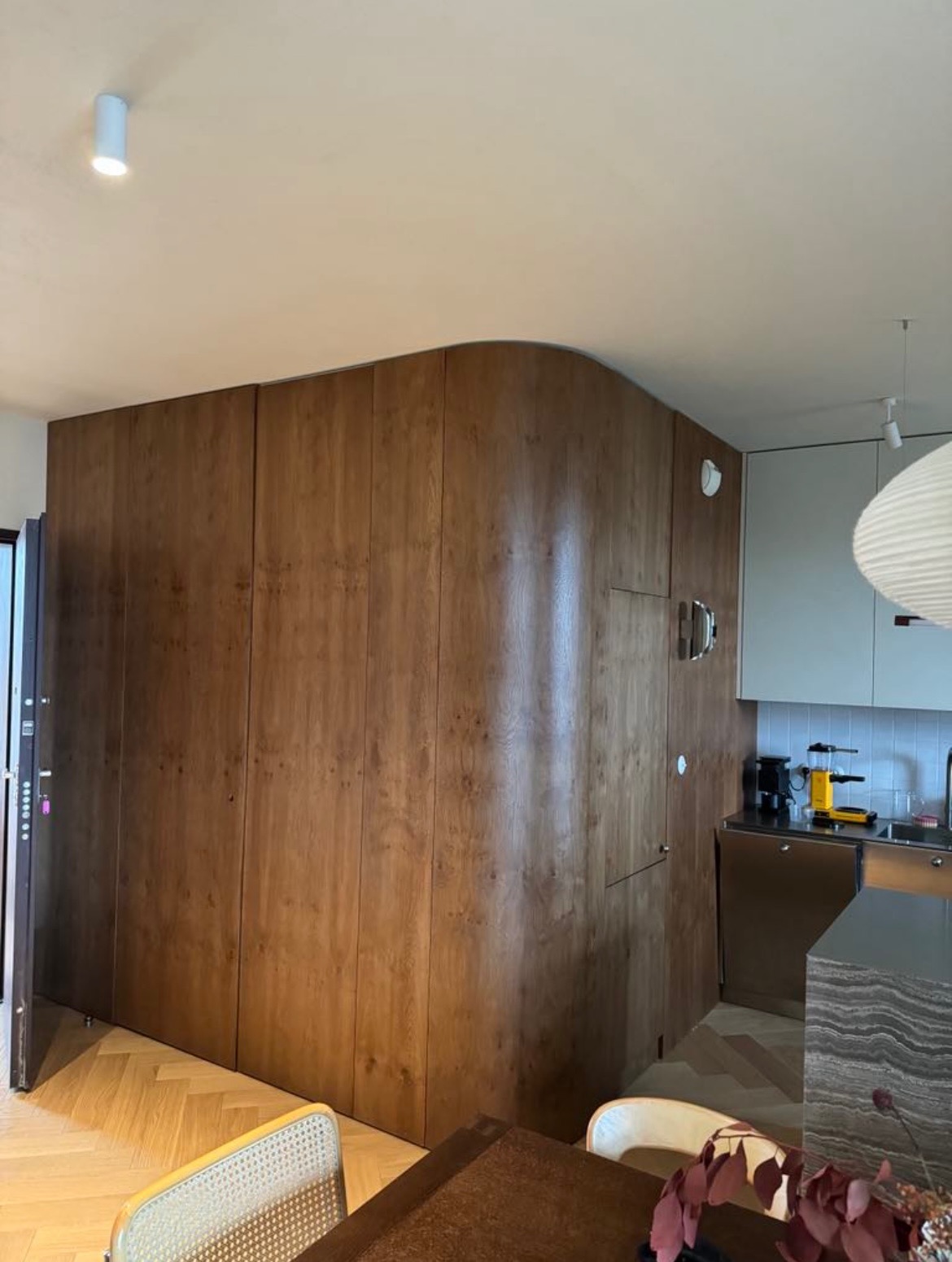 Drewniana szafa wnękowa z zaokrąglonym narożnikiem, wkomponowana w przestrzeń kuchni. Widoczne ukryte drzwi i minimalizm.