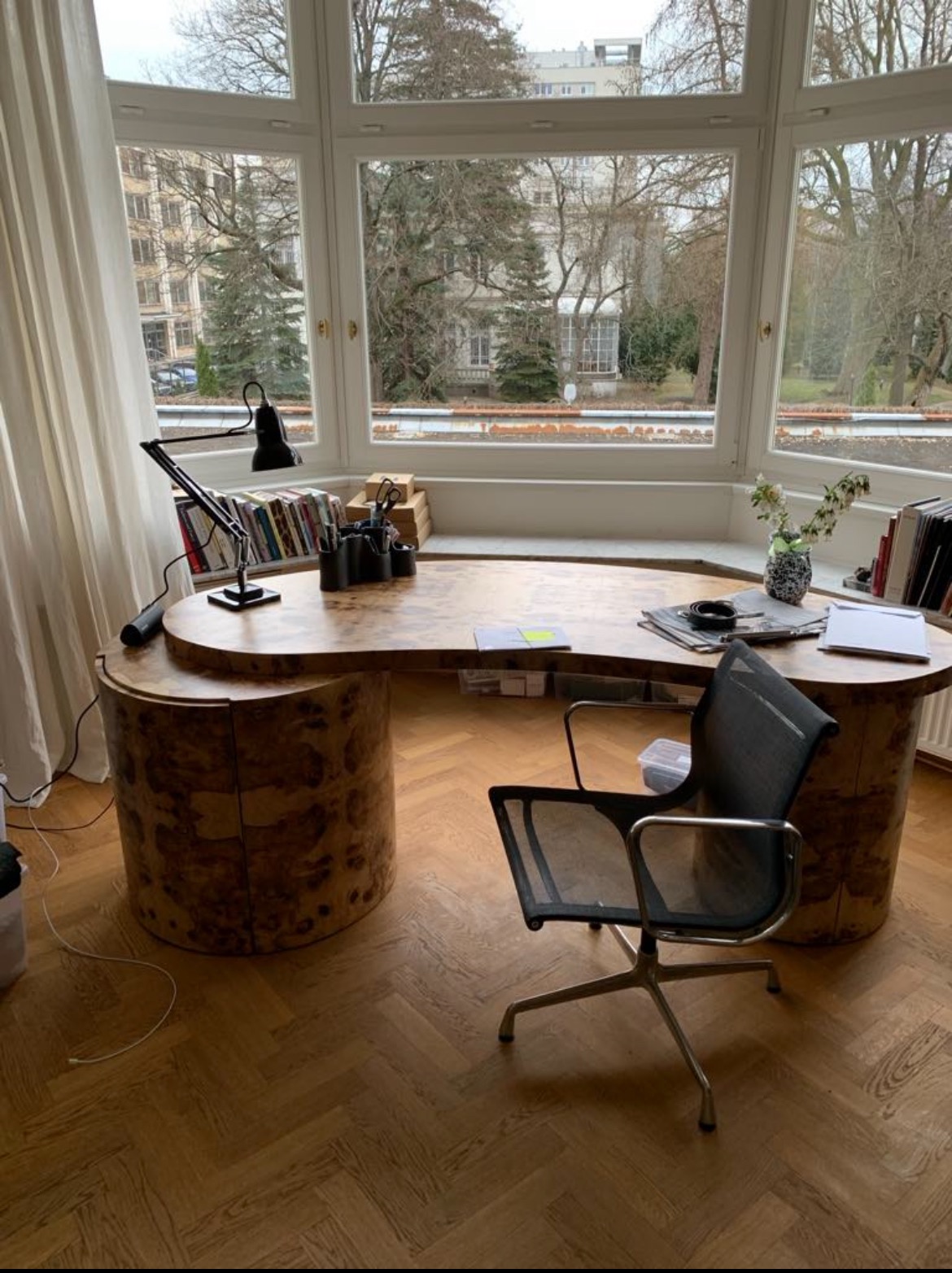 Biurko z drewna o nieregularnym kształcie, z widoczną strukturą słojów, stoi w pokoju z dużym oknem i parkietem w jodełkę. Krzesło biurowe obok.