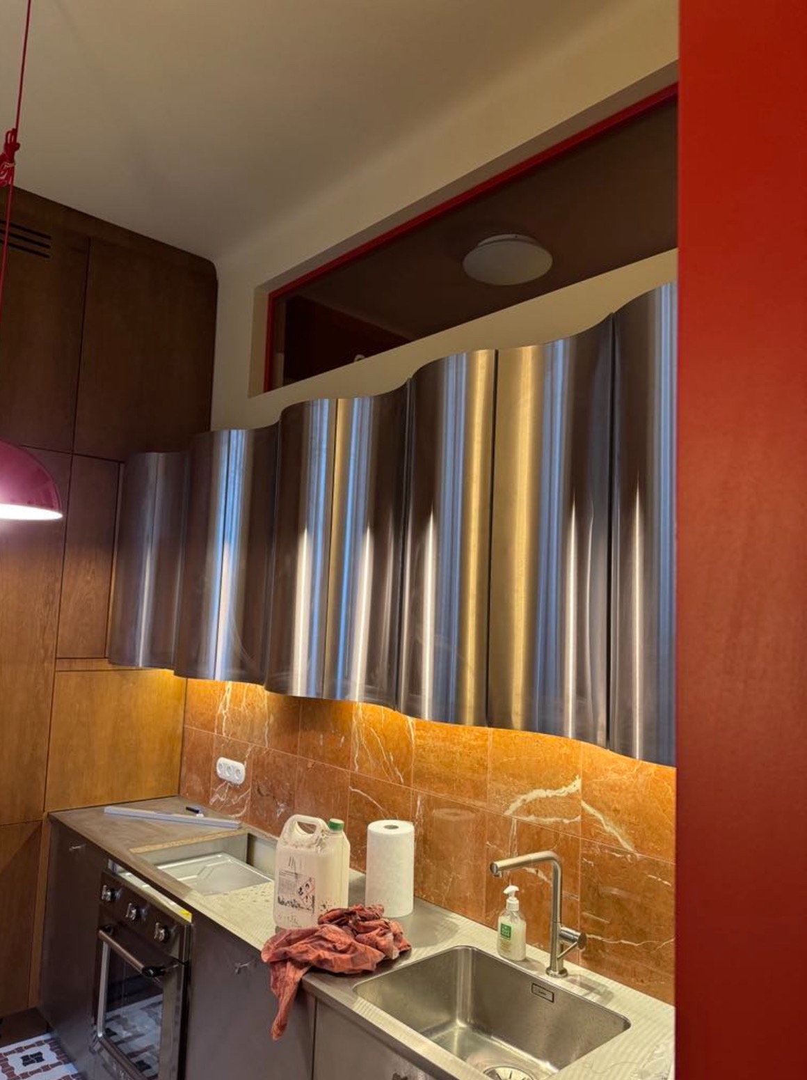 Nowoczesna kuchnia z falującymi, metalowymi szafkami wiszącymi i pomarańczowymi płytkami marmurowymi. Widoczne akcesoria kuchenne na blacie.