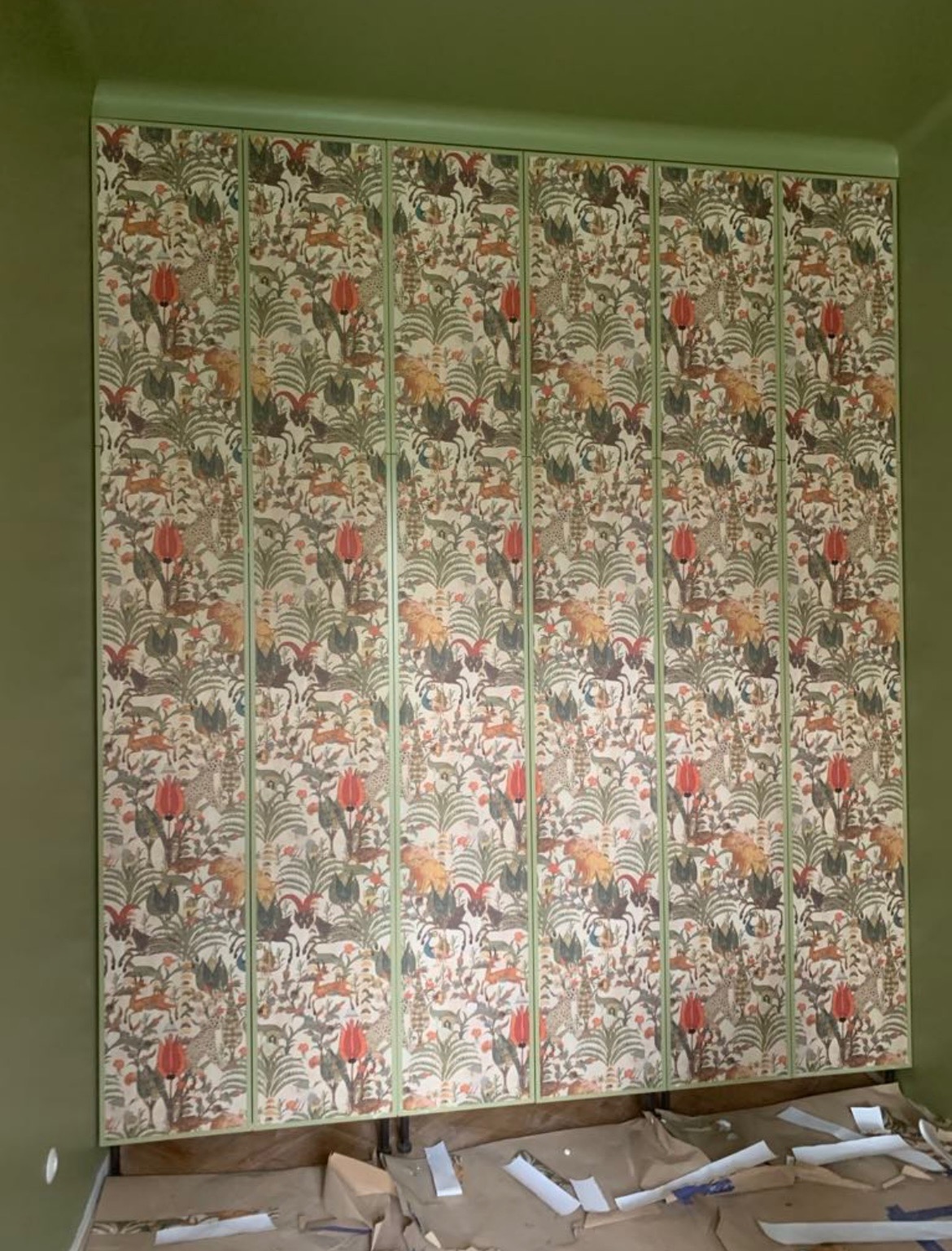 Szafa wnękowa z ozdobnymi frontami w stylu vintage, dopasowana kolorystycznie do zielonych ścian. Widoczne zabezpieczenia podłogi przed zabrudzeniem.