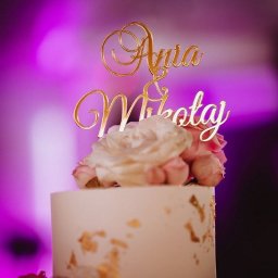 WED IT WEDDINGS KATARZYNA WÓJCIAK - Elegancki tort weselny z białym lukrem, złotymi płatkami, kwiatami i topperem z imionami 'Ania & Mikołaj' na tle fioletowego oświetlenia.