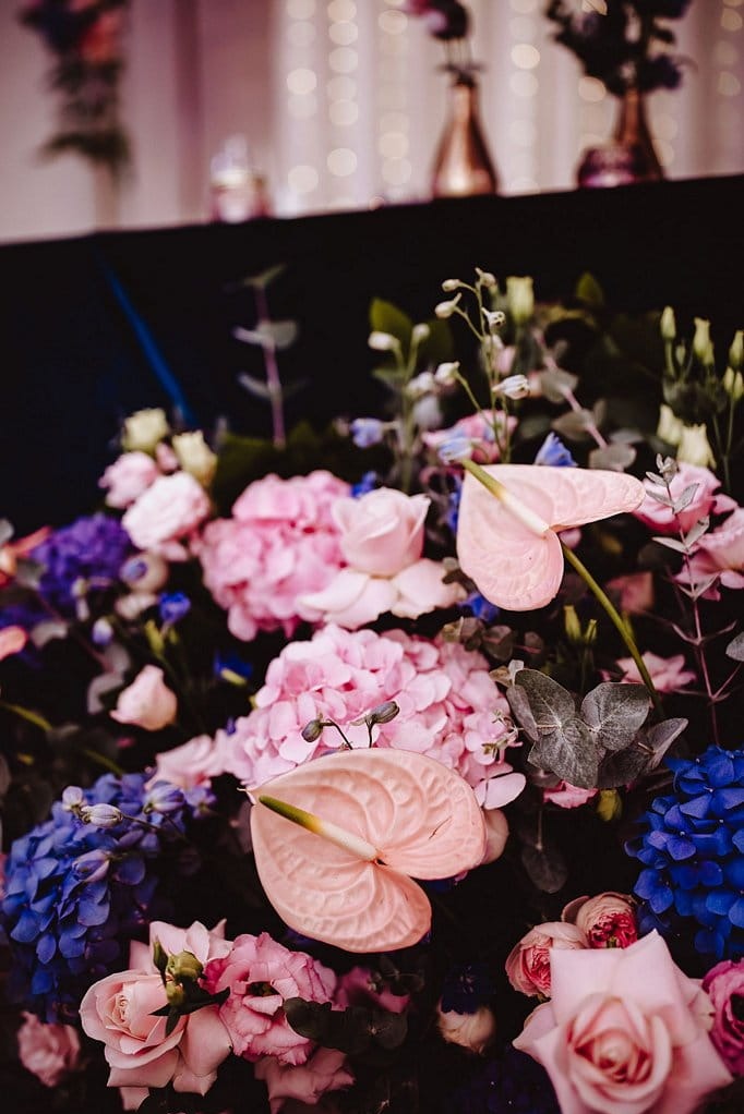 Kompozycja kwiatowa z róż, hortensji i anturium w odcieniach różu i fioletu na ciemnym tle, dekoracja stołu z elementami oświetlenia w tle.