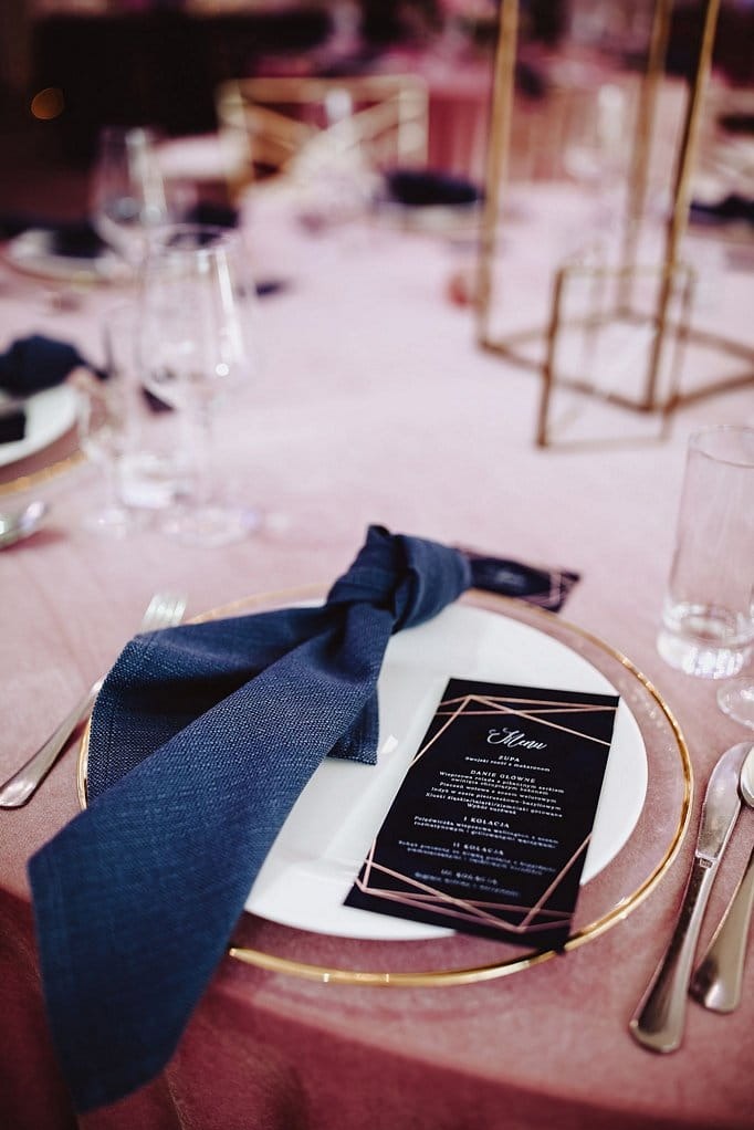 Elegancko nakryty stół z menu i granatową serwetką na białym talerzu ze złotym obramowaniem, różowy obrus, szklanki i sztućce w tle.