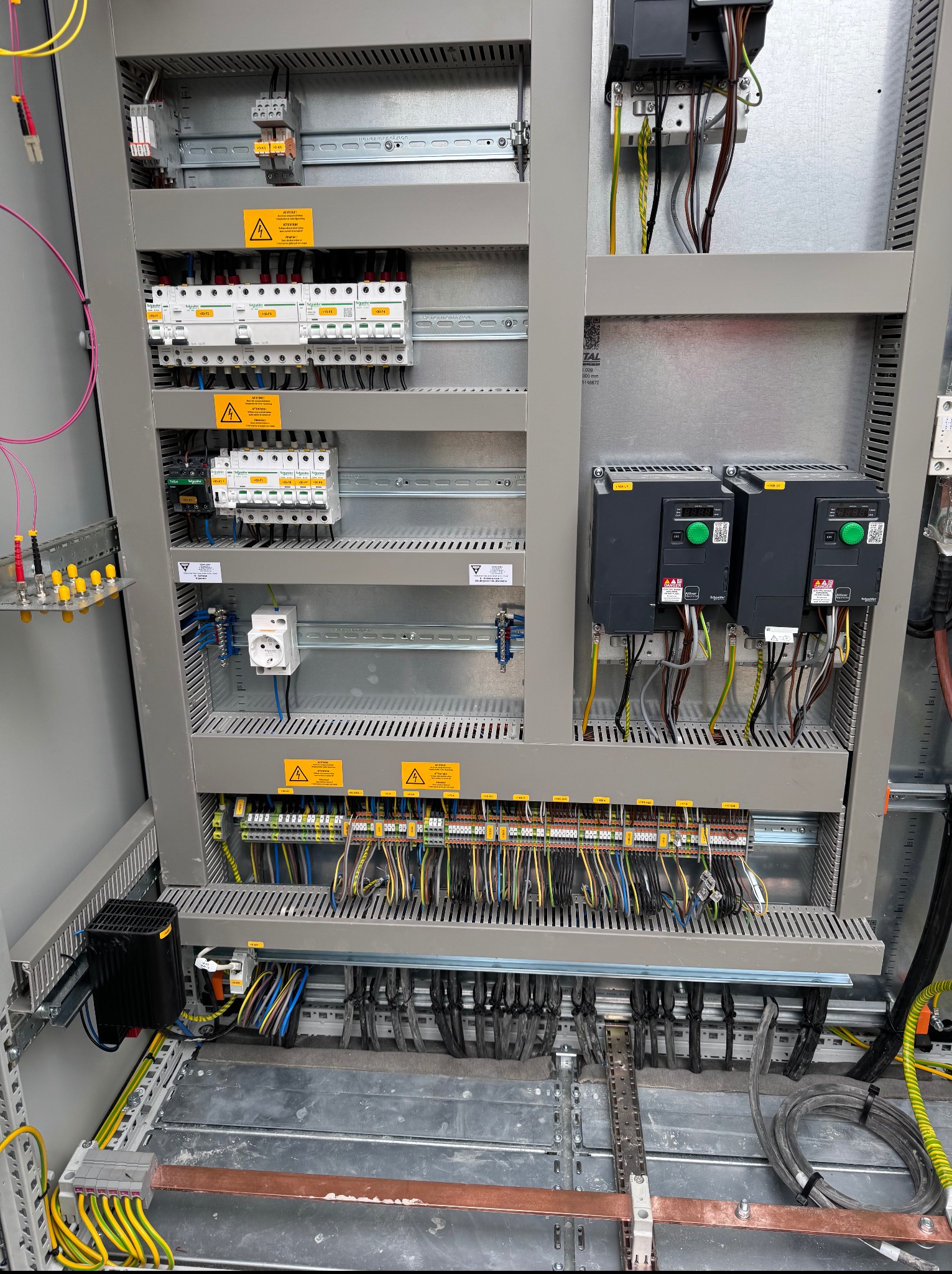 Szczegółowe ujęcie wnętrza szafy elektrycznej z licznymi przewodami, bezpiecznikami i elementami sterującymi. Widoczne żółte oznaczenia ostrzegawcze.