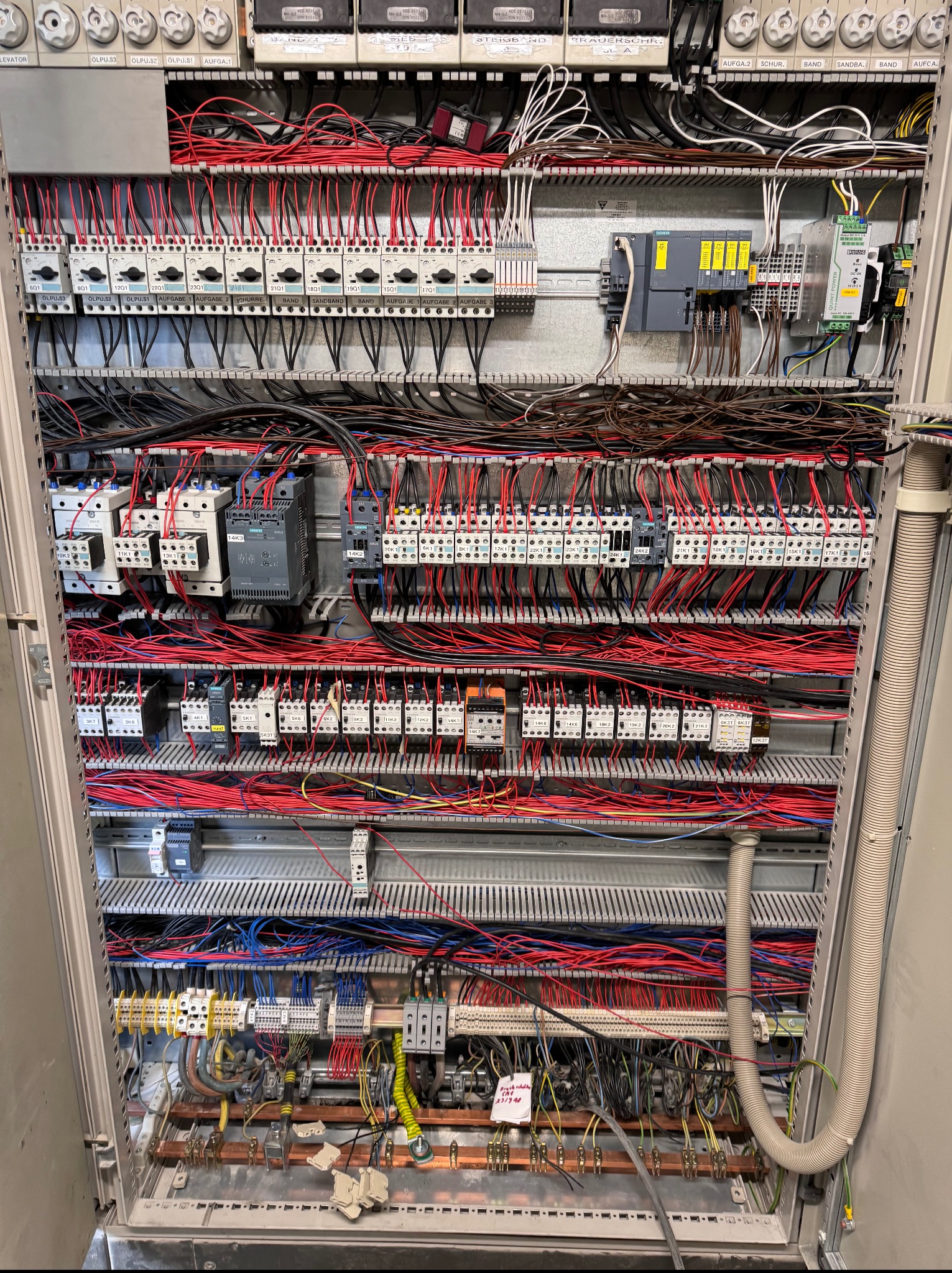 Rozbudowana szafa sterownicza z licznymi przewodami, stycznikami i zabezpieczeniami, widoczne oznaczenia na elementach, uporządkowany układ przewodów w kolorach czerwonym i czarnym.