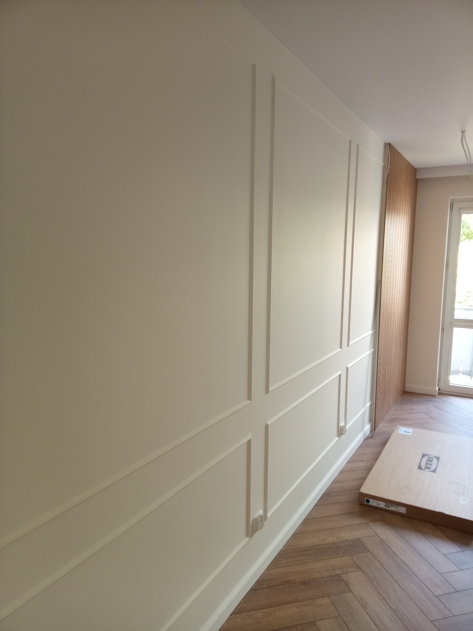 Ściana z dekoracyjnymi panelami w odcieniach bieli, parkiet w jodełkę i fragment drewnianej ściany. Na podłodze kartonowe opakowanie.