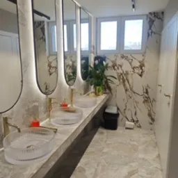 Elegancka łazienka z marmurowymi ścianami, trzema umywalkami z przezroczystego szkła i złotymi bateriami. Lustra z podświetleniem LED i rośliny dodają luksusowego charakteru.