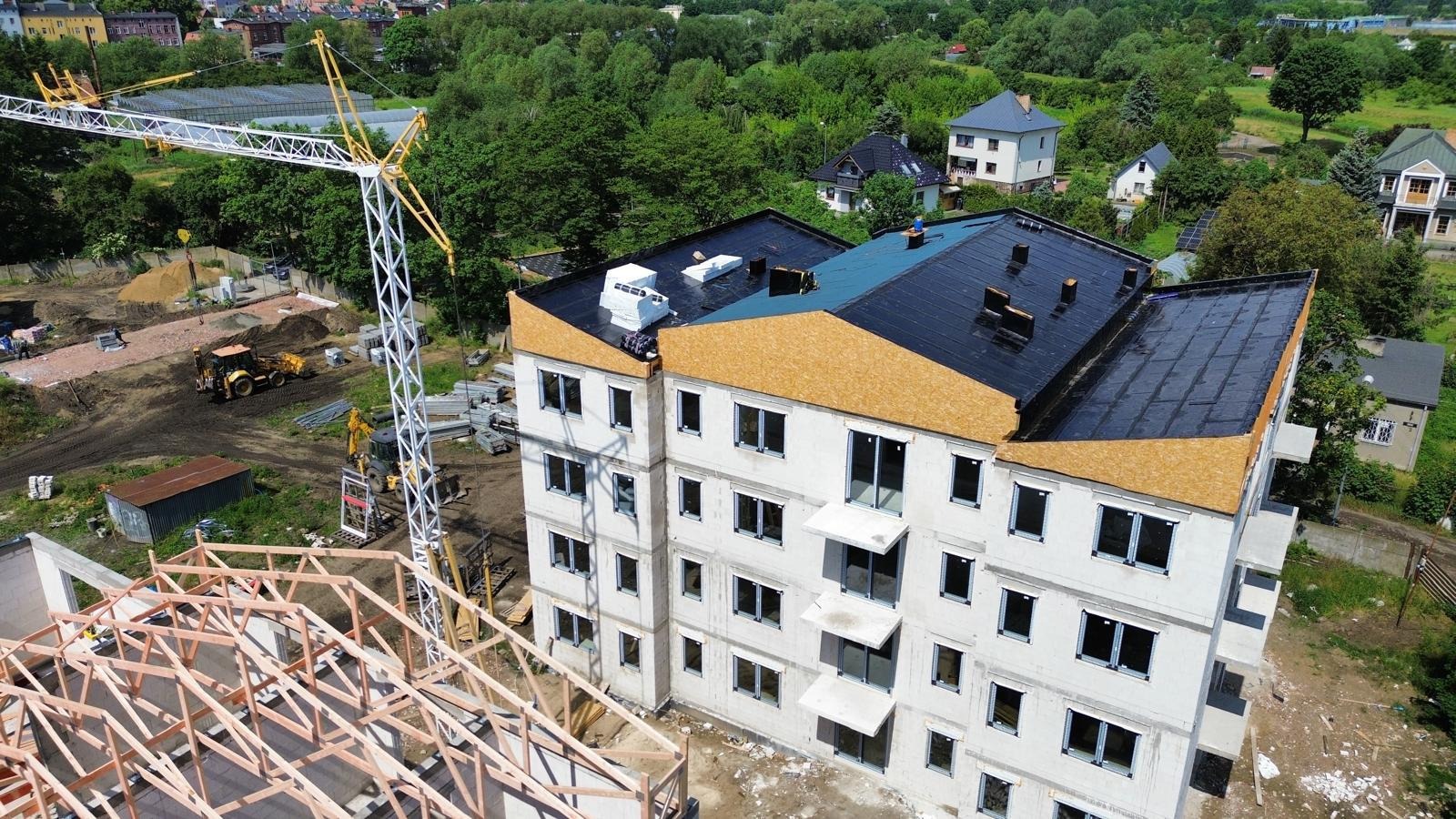 Wysoki budynek w stanie surowym zamkniętym z czarnym dachem i oklejonymi płytami OSB poddaszem, widziany z góry w słoneczny dzień. Na pierwszym planie szkielet konstrukcji.