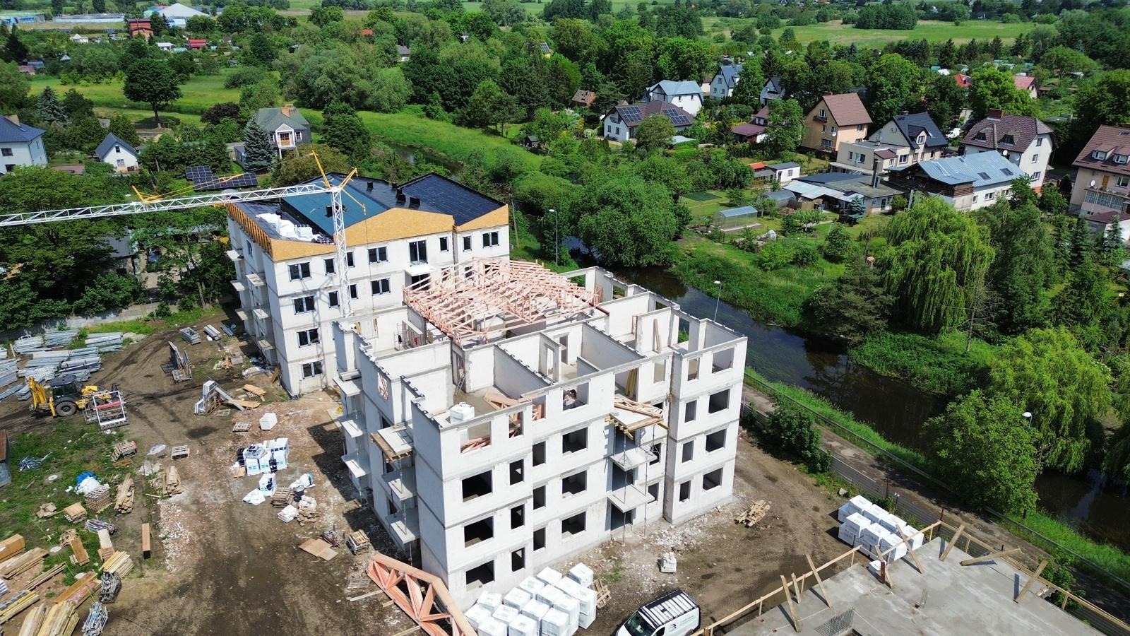 Budowa bloku mieszkalnego w stanie surowym otoczonego zielenią, widok z góry. Częściowo widoczne elementy konstrukcji dachu i elementy wykończeniowe.