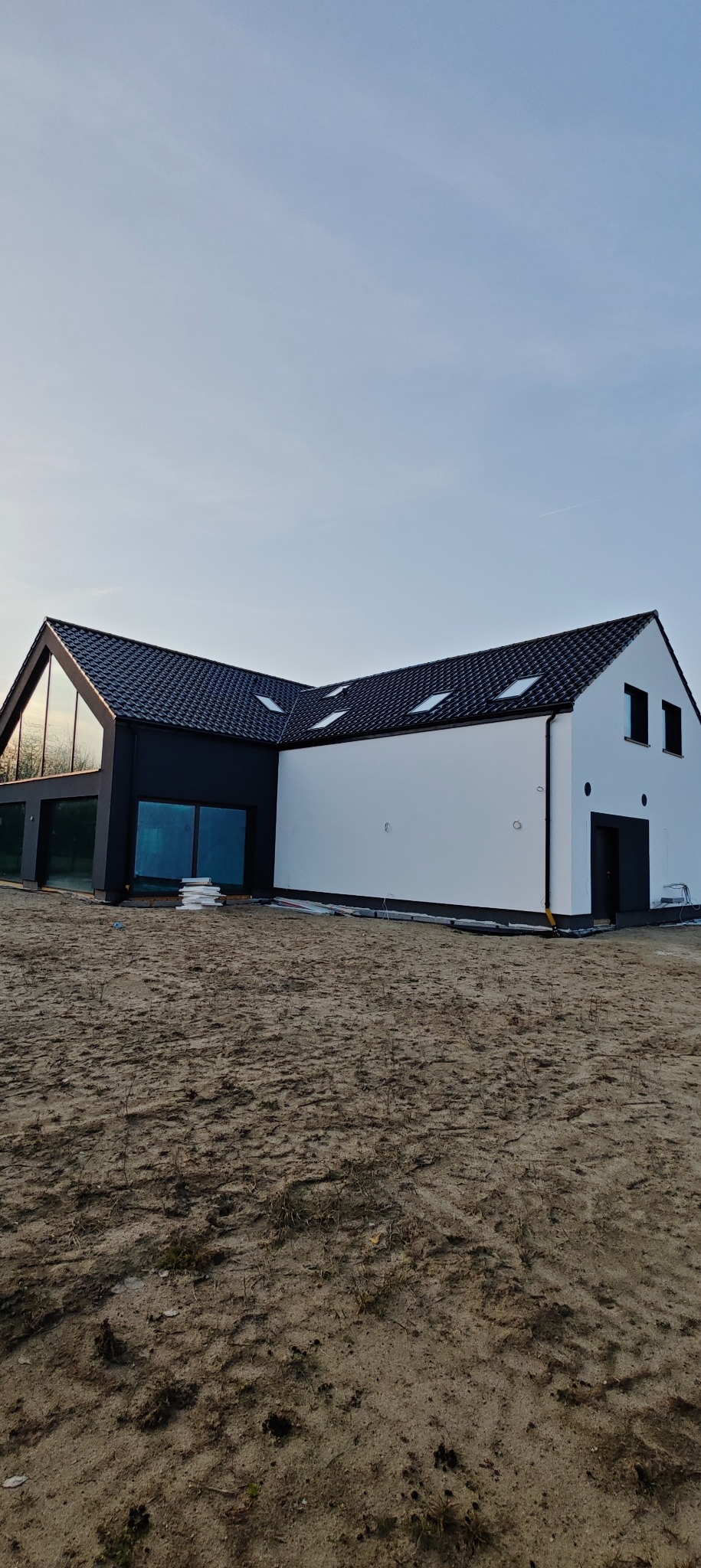 Nowoczesny dom z czarno-białą elewacją i dużymi oknami, otoczony piaszczystym terenem. Widoczne dachówki i fragmenty instalacji elektrycznej przy budynku.