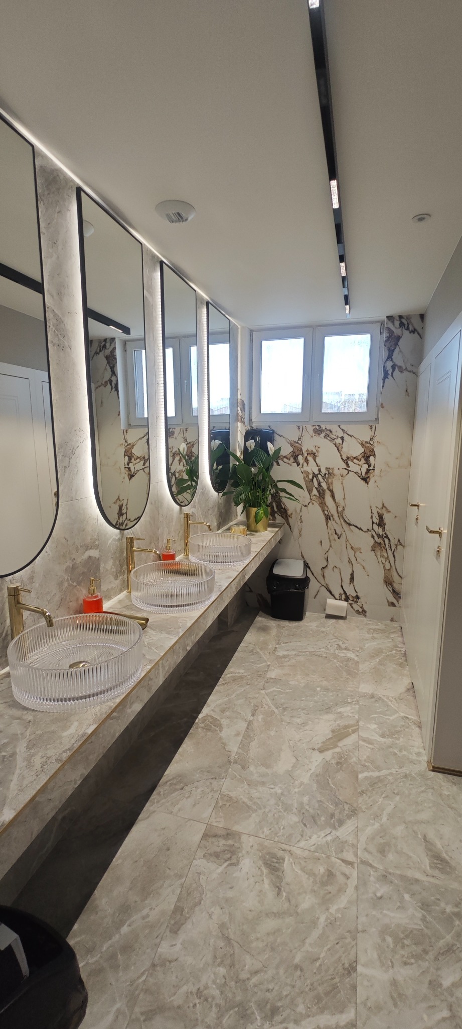 Elegancka łazienka z marmurowymi ścianami, trzema umywalkami z przezroczystego szkła i złotymi bateriami. Lustra z podświetleniem LED i rośliny dodają luksusowego charakteru.