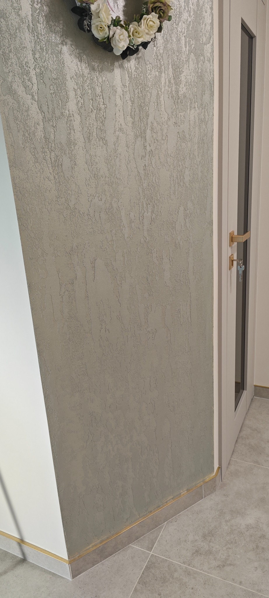 Dekoracyjna ściana z efektem trawertynu w odcieniach szarości, listwa przypodłogowa w kolorze złota, białe drzwi z przeszkleniem i wieniec z kwiatów.