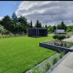 Zadbany trawnik z r&oacute;wno przystrzyżoną trawą i rabatami kwiatowymi, altana ogrodowa w tle, widoczny samoch&oacute;d dostawczy. Słoneczny dzień, częściowo zachmurzone niebo.