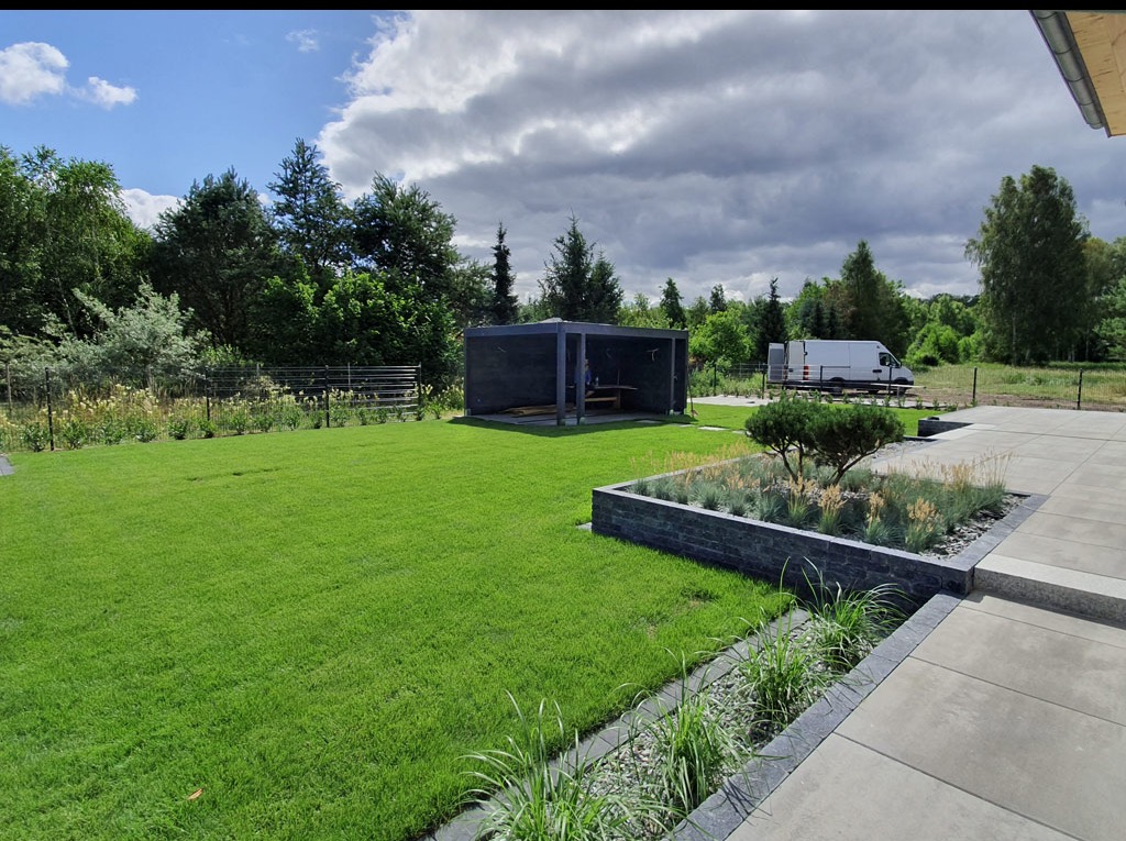 Zadbany trawnik z równo przystrzyżoną trawą i rabatami kwiatowymi, altana ogrodowa w tle, widoczny samochód dostawczy. Słoneczny dzień, częściowo zachmurzone niebo.
