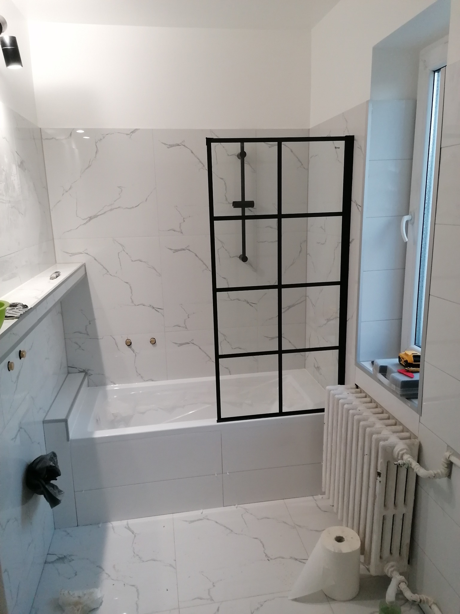 Nowoczesna aranżacja łazienki w Bydgoszczy: białe płytki imitujące marmur, czarna kabina prysznicowa typu loft i grzejnik żeberkowy. Minimalistyczny design.