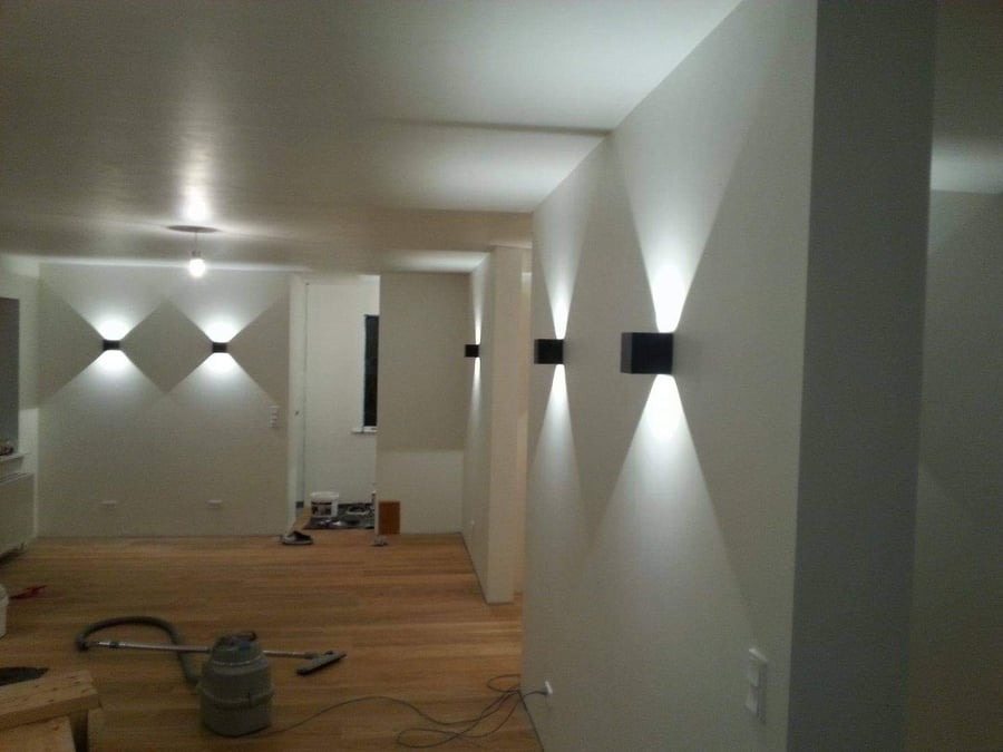 Nowoczesne oświetlenie ścienne z czarnymi lampami emitującymi snopy światła na gładkich, białych ścianach w odnowionym wnętrzu z parkietem i widocznym odkurzaczem.