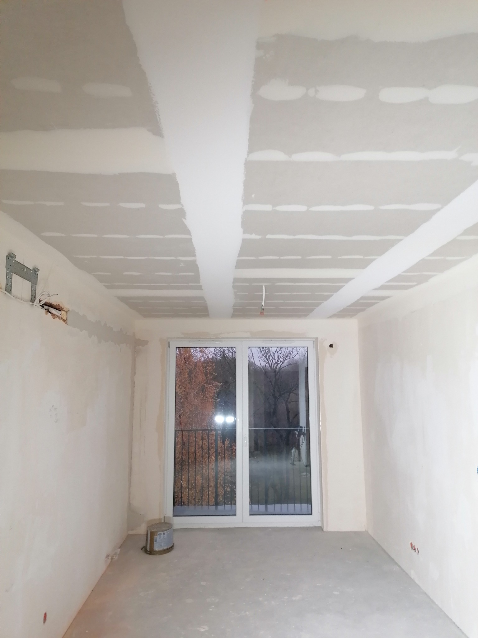 Pokój w trakcie prac wykończeniowych: szpachlowane ściany i sufit z płyt karton-gips, widoczne łączenia. Okno balkonowe z widokiem na zewnątrz.