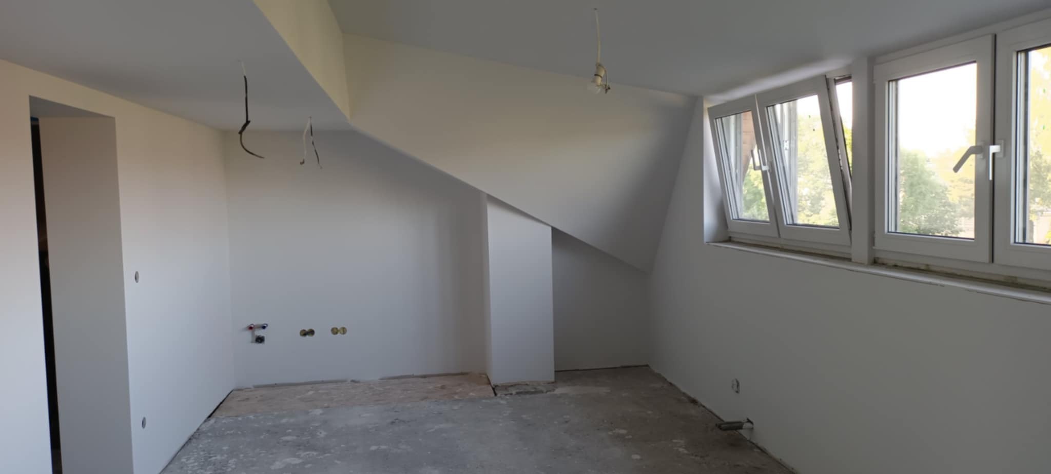Wnętrze pokoju na poddaszu po szpachlowaniu i malowaniu ścian na biało. Widoczne okna, instalacje i surowa podłoga przed wykończeniem.
