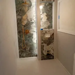 Wykończona łazienka z marmurową ścianą prysznicową z złotą armaturą i podtynkową toaletą. Widoczne białe płytki podłogowe i fragment instalacji elektrycznej.