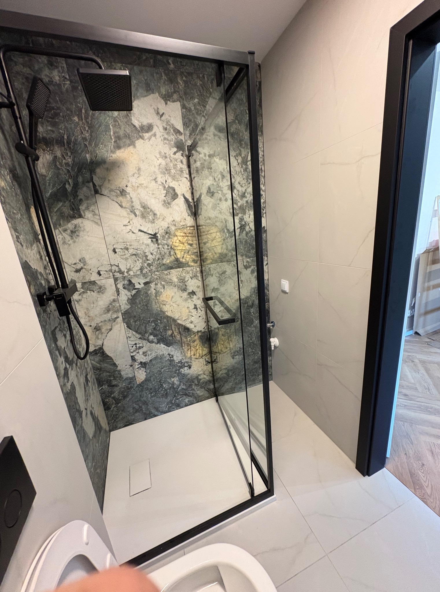 Nowoczesna kabina prysznicowa z czarnymi ramami i deszczownicą na tle marmurowych płytek, widok z toalety. Minimalistyczny design i eleganckie wykończenie.