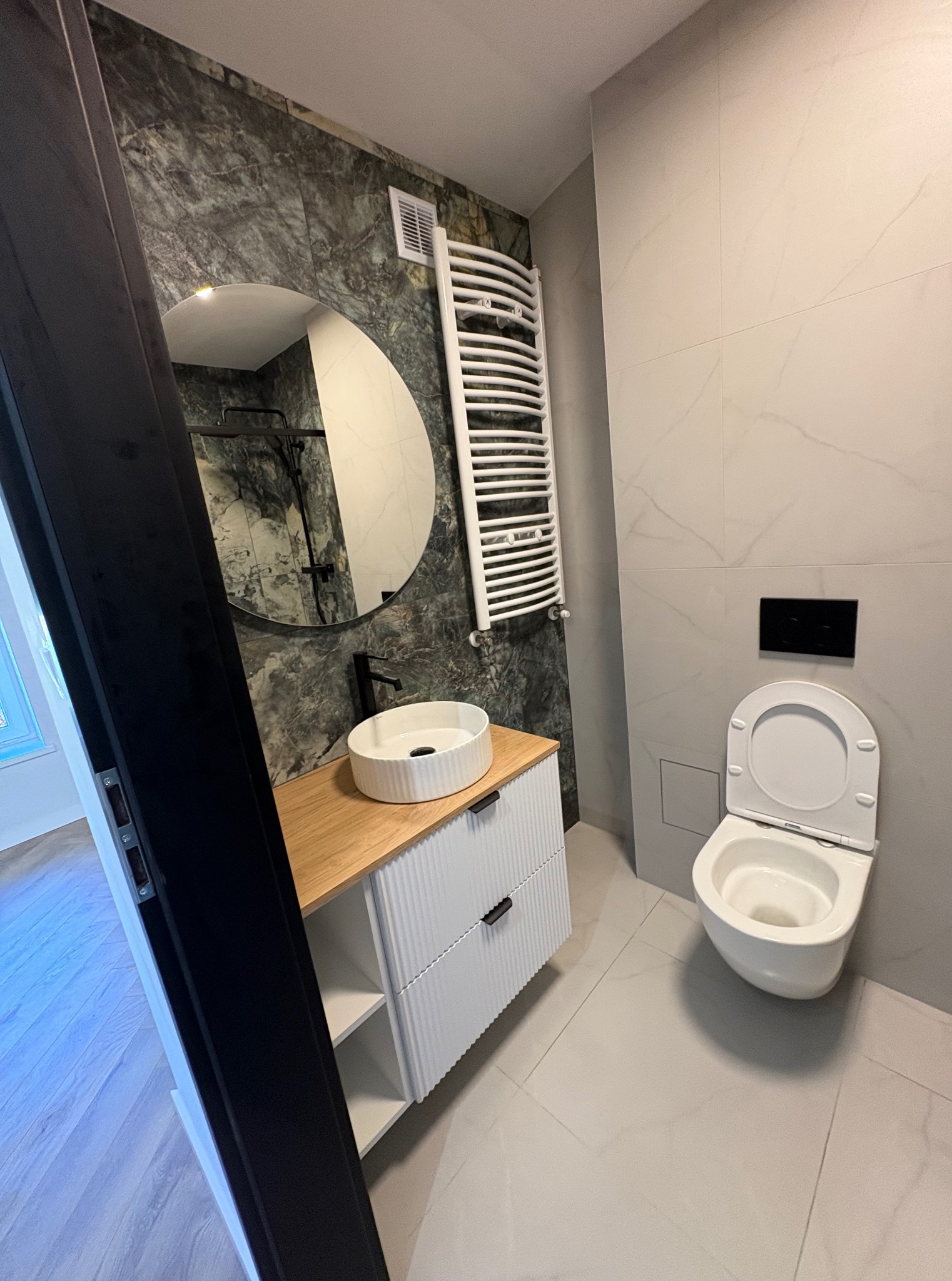 Nowoczesna łazienka z umywalką nablatową, okrągłym lustrem i grzejnikiem drabinkowym. Marmurowe płytki na ścianach, minimalistyczna toaleta. Widok zza otwartych drzwi.