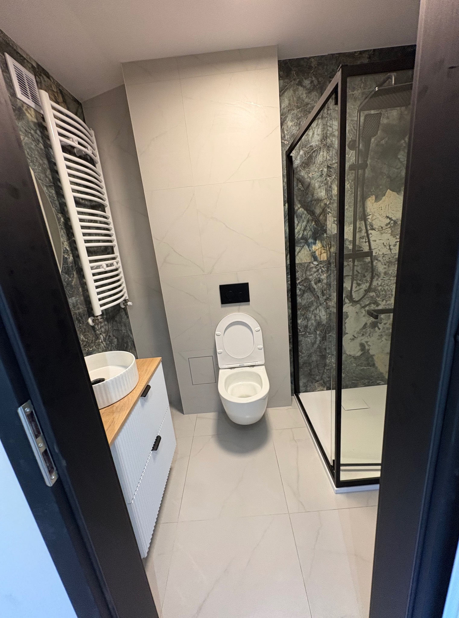 Nowoczesna łazienka z kabiną prysznicową w czarnej ramie, umywalką nablatową i grzejnikiem drabinkowym. Marmurowe płytki na ścianach i podłodze. Widok zza czarnej futryny.