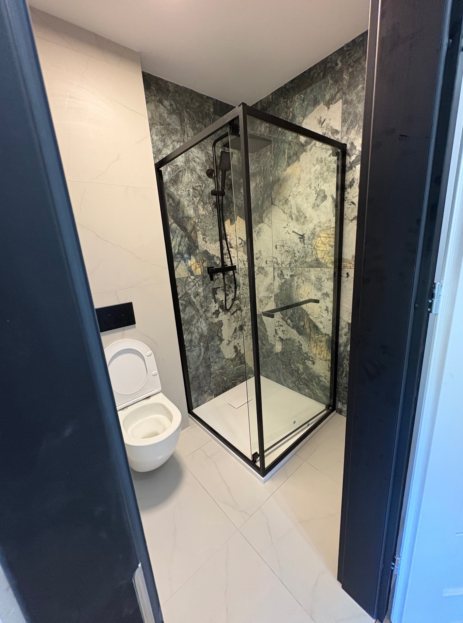 Nowoczesna łazienka z kabiną prysznicową w czarnej ramie i toaletą podwieszaną. Marmurowe płytki na ścianach, biała podłoga. Widok zza framugi drzwi.