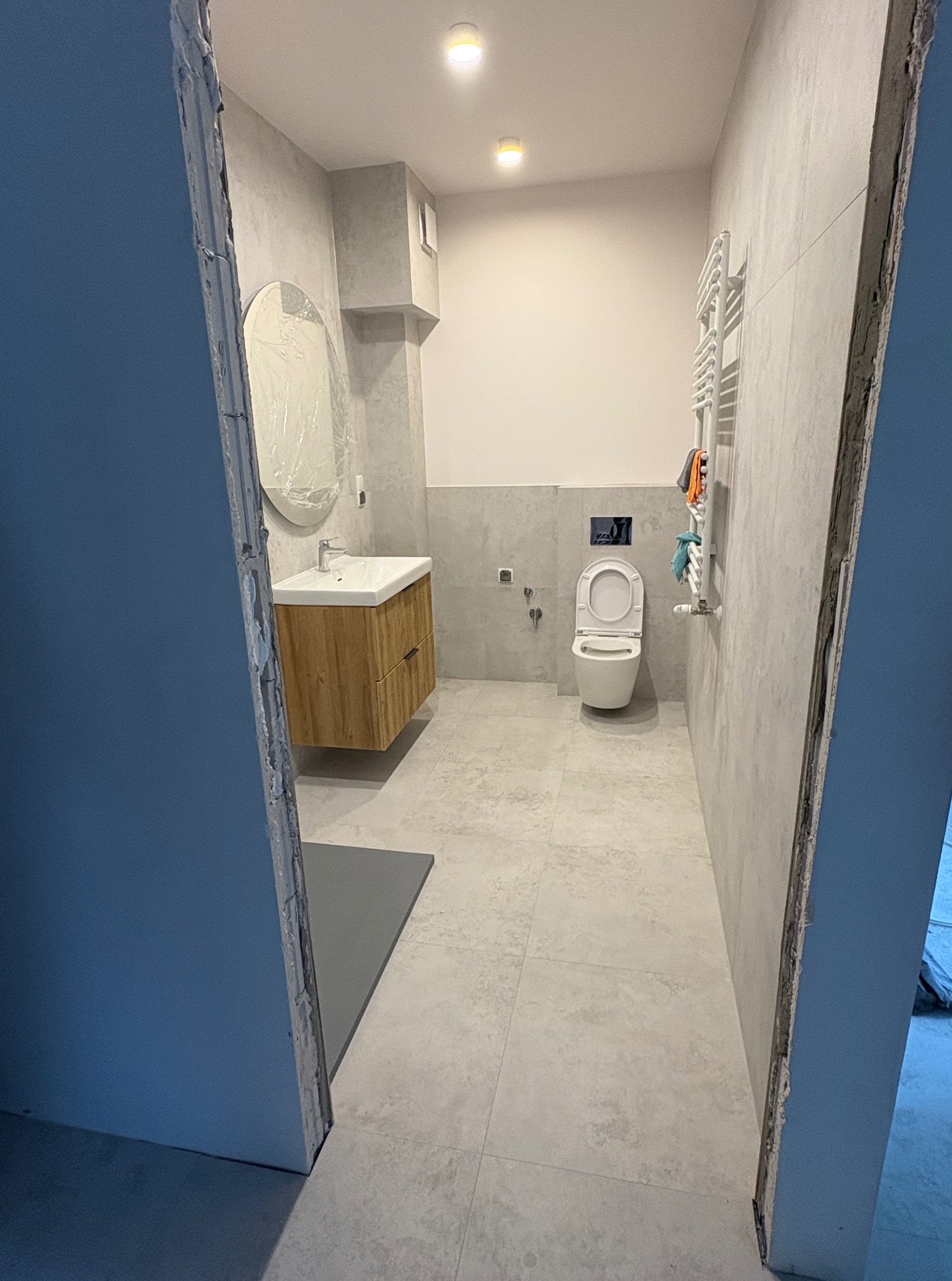 Wykończona łazienka w szarościach z drewnianą szafką pod umywalką i grzejnikiem drabinkowym. Ujęcie zza framugi, owalne lustro w folii ochronnej.