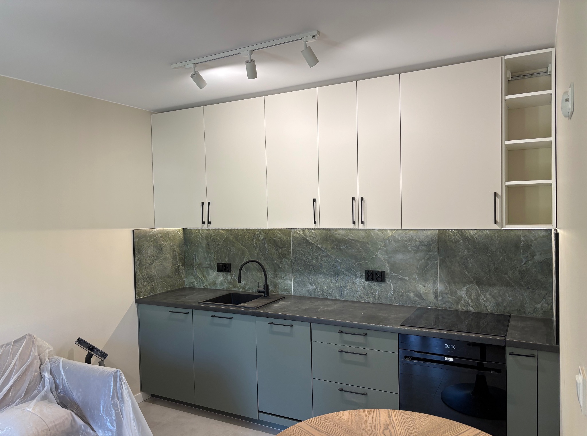 Nowoczesna kuchnia z zielonymi szafkami i marmurowym backsplashiem. Białe szafki wiszące z czarnymi uchwytami, oświetlenie punktowe i sprzęt AGD w zabudowie.