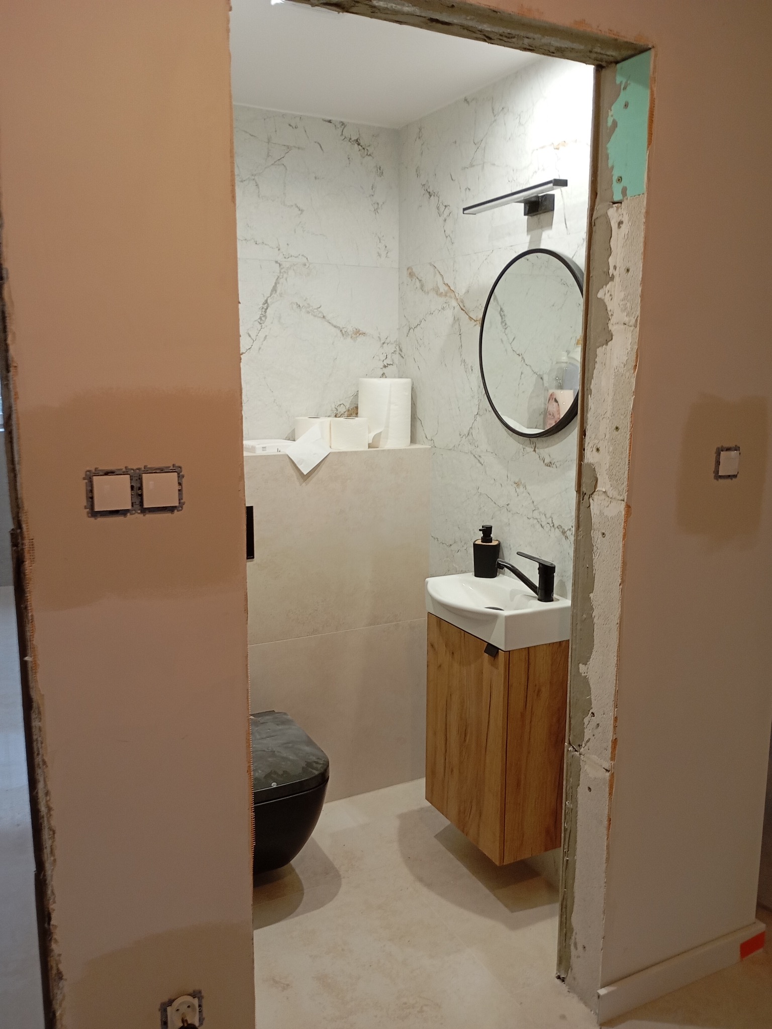 Wnętrze łazienki w trakcie wykańczania: widok na umywalkę z szafką, lustro, toaletę i marmurowe płytki. Surowe ściany i otwory na gniazdka elektryczne.