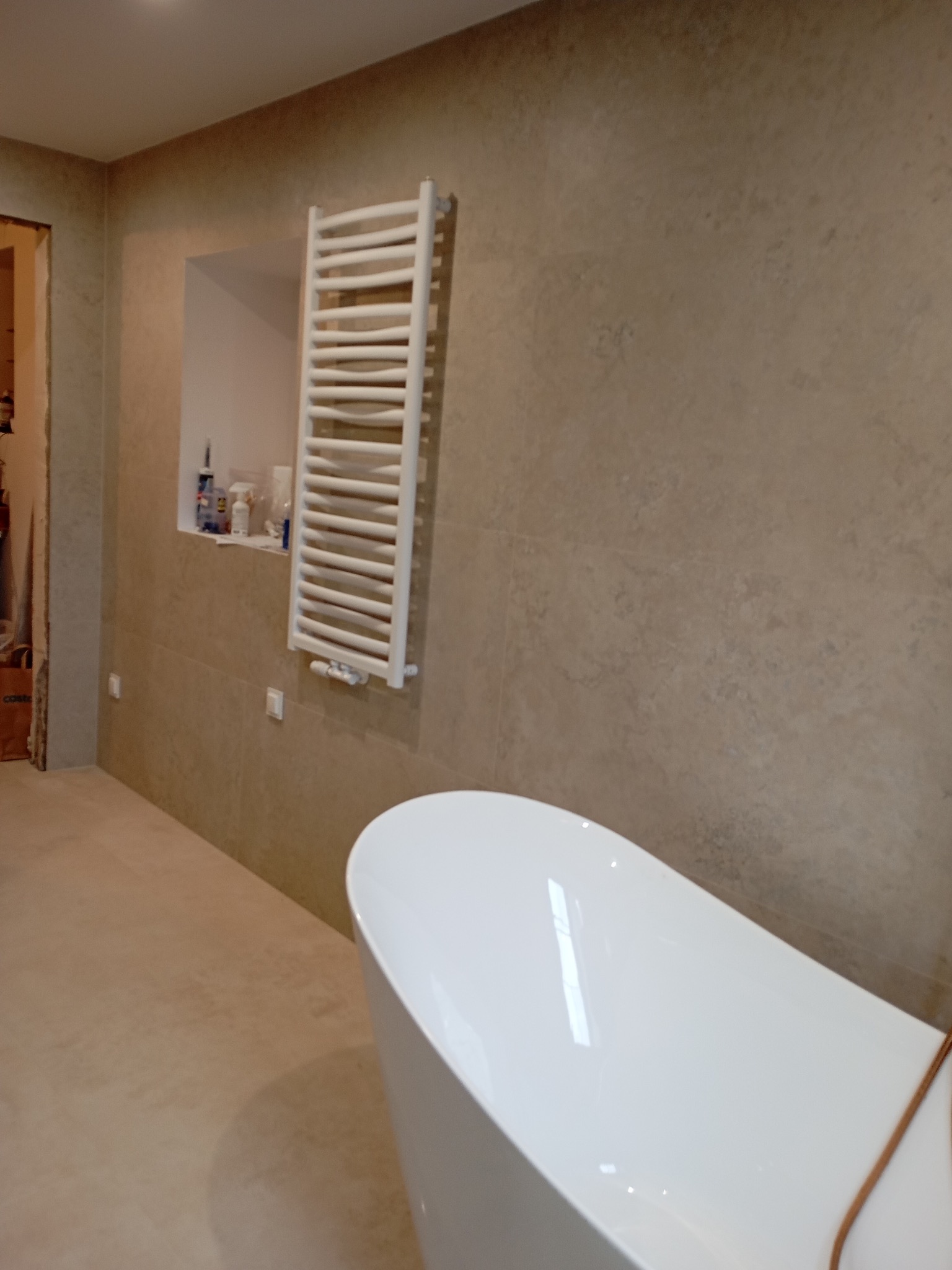 Nowoczesna łazienka z beżowymi płytkami, białą wanną wolnostojącą i grzejnikiem drabinkowym. W ścianie wnęka z kosmetykami. Ukończone wykończenie wnętrza.