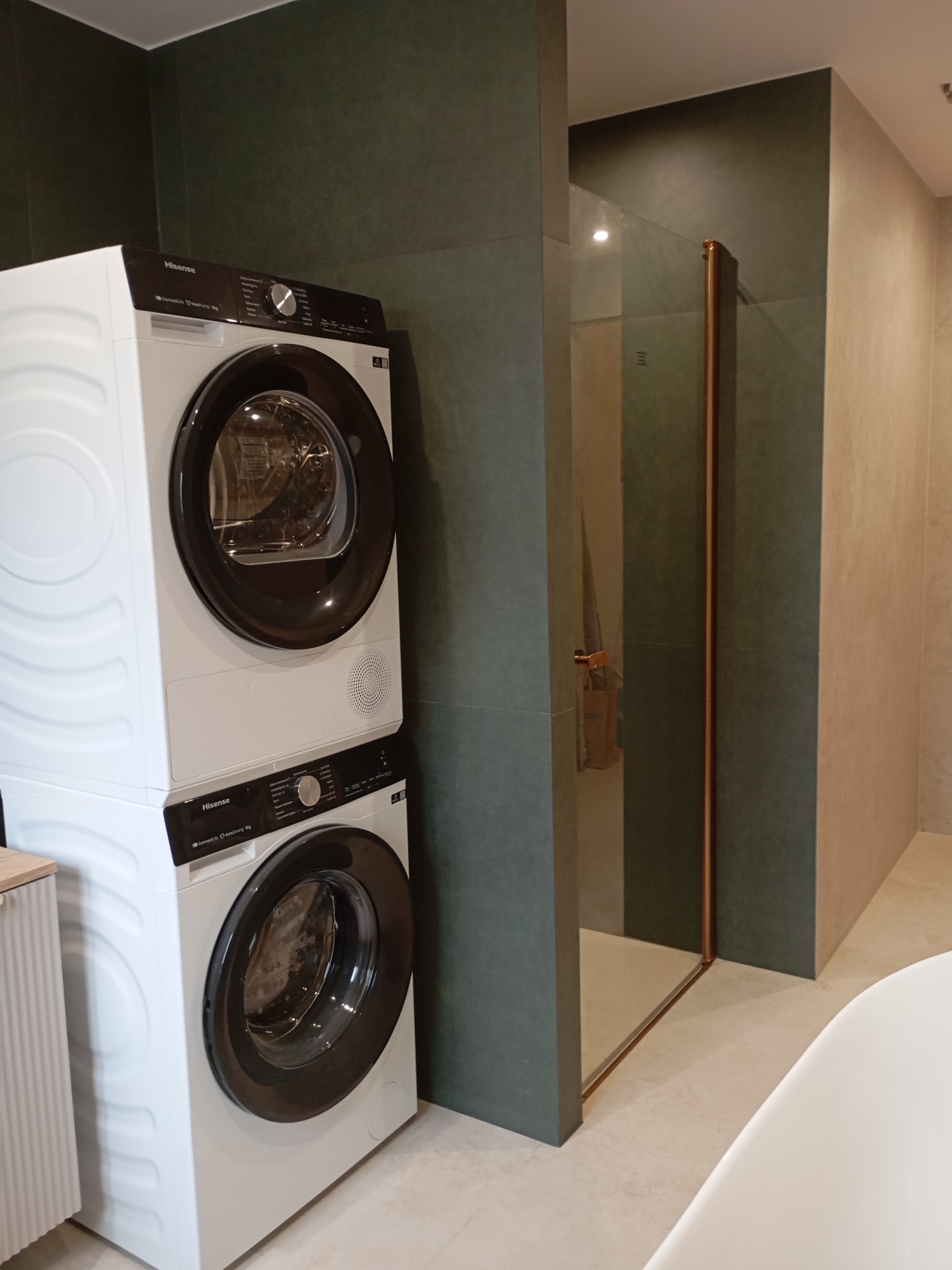 Nowoczesna łazienka z pralko-suszarką Hisense w zabudowie i kabiną prysznicową z miedzianymi profilami na tle ciemnozielonych płytek.