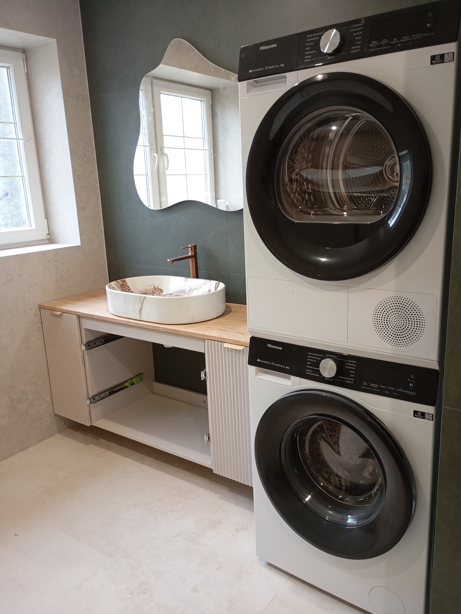 Nowoczesna łazienka z umywalką nablatową, szafką z otwartymi szufladami i pralko-suszarką Hisense w pionie. Nietypowe lustro i stonowana kolorystyka.