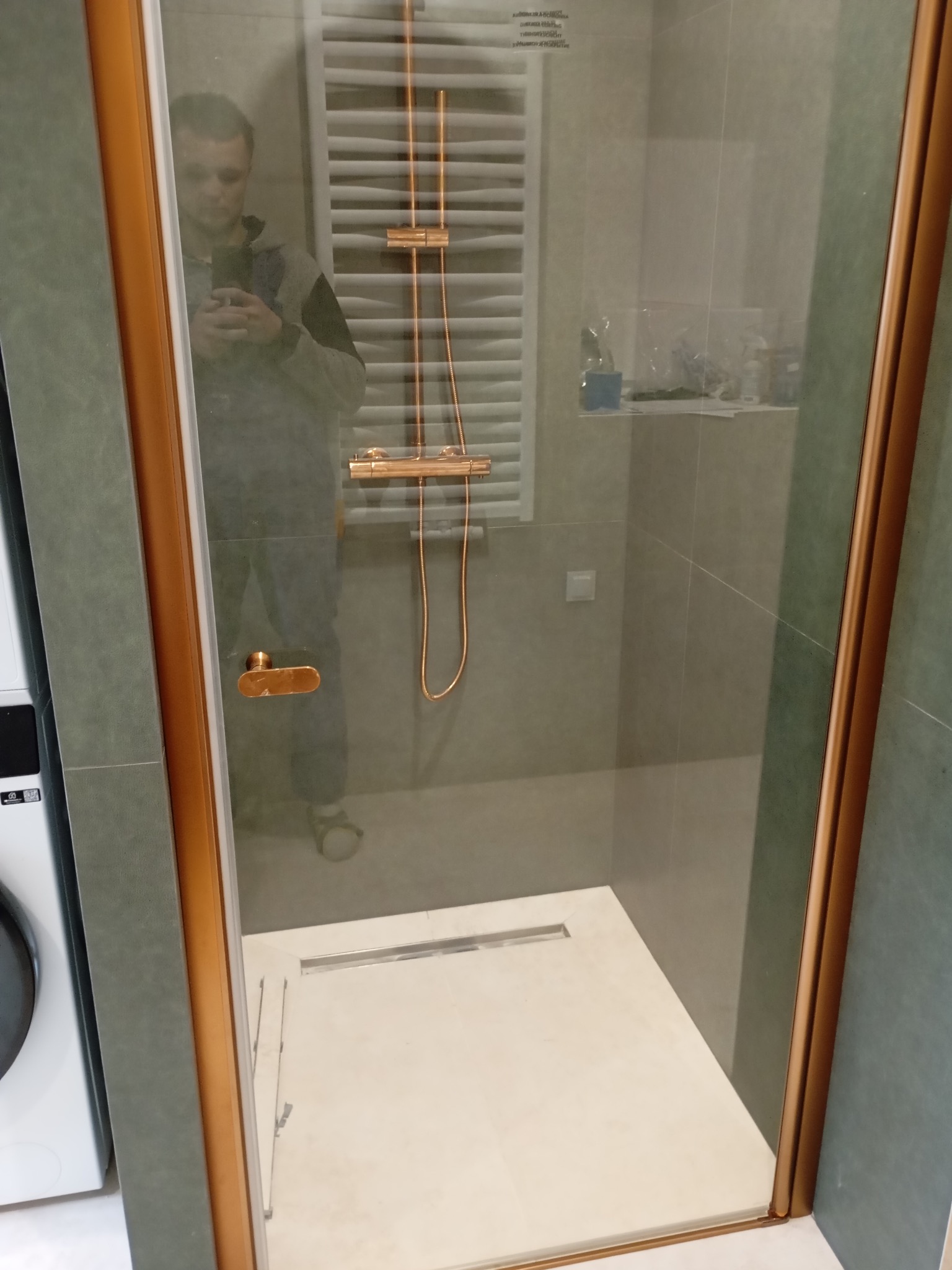 Nowoczesna kabina prysznicowa z elementami w kolorze miedzi, widoczna w łazience po remoncie w Jeleniej Górze. Uwagę zwraca odpływ liniowy i grzejnik drabinkowy.