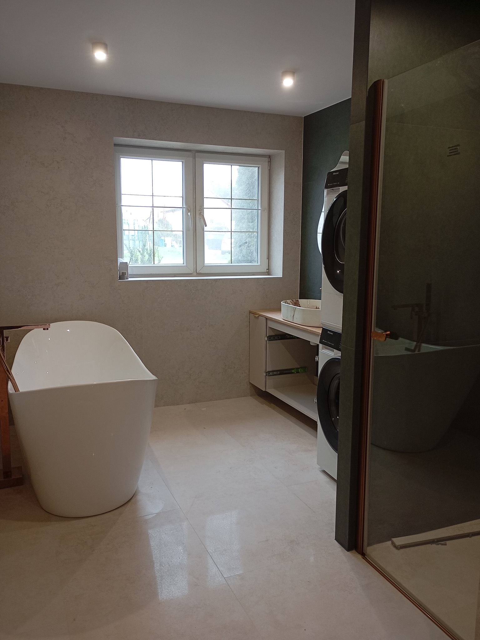 Nowoczesna łazienka z wolnostojącą wanną, oknem i pralko-suszarką w zabudowie. Beżowe płytki, minimalistyczny design.