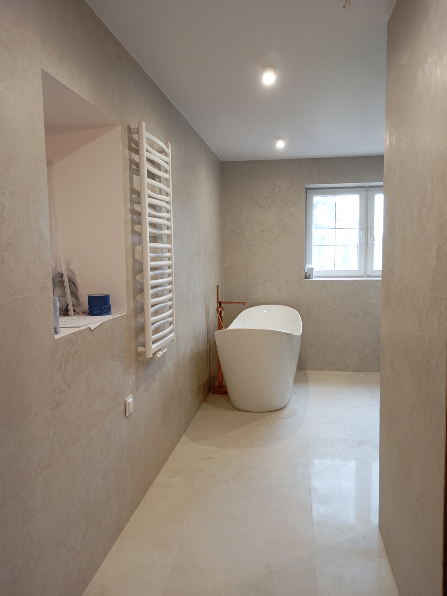 Nowoczesna łazienka z wolnostojącą wanną, grzejnikiem drabinkowym i beżowymi płytkami. W oknie widoczne światło dzienne. Minimalistyczny design.