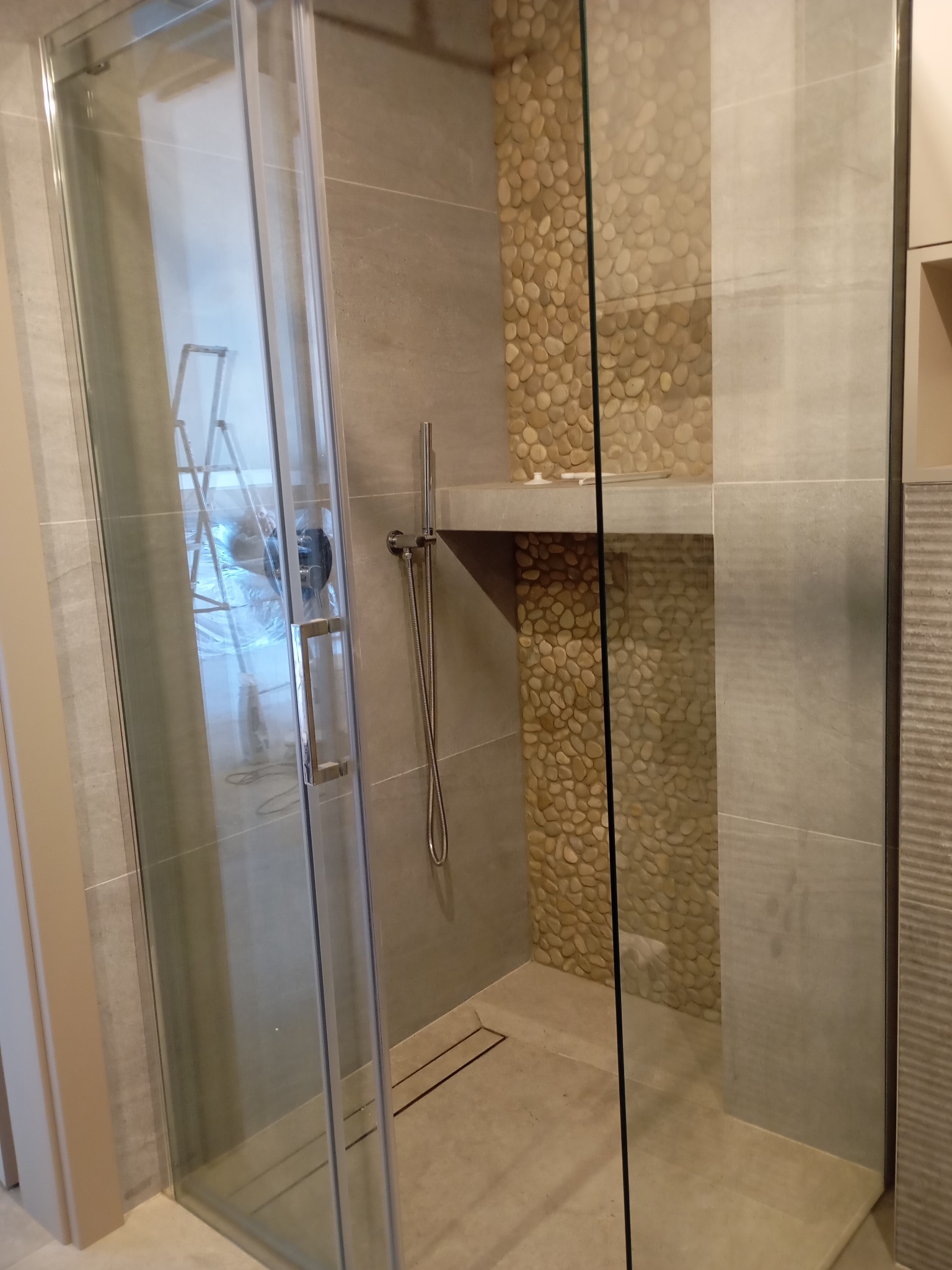 Nowoczesna kabina prysznicowa z szarymi płytkami i dekoracyjną ścianą z kamieni rzecznych. Widoczna drabina malarska w odbiciu szyby.