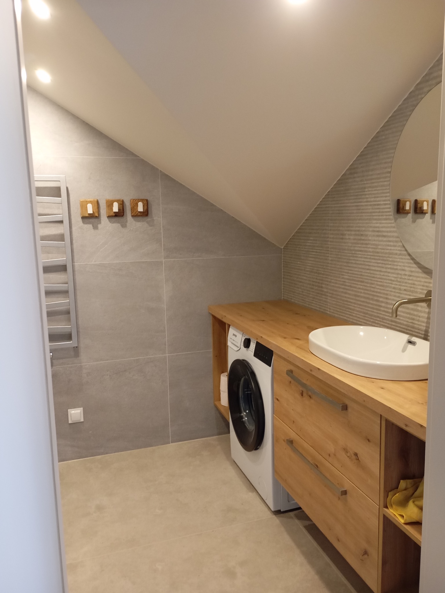 Nowoczesna łazienka na poddaszu z drewnianą zabudową pralki, umywalką nablatową i szarą glazurą. Oświetlenie punktowe podkreśla skosy.