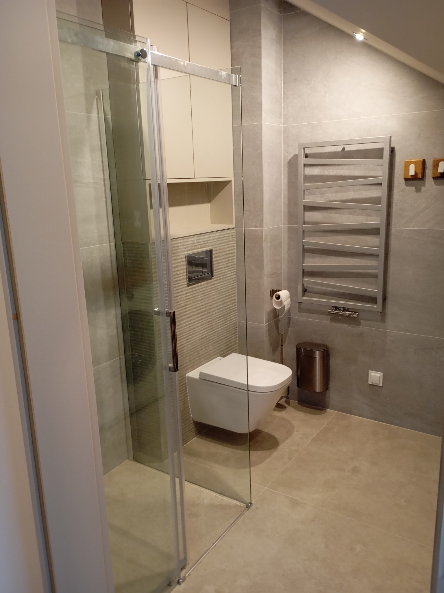 Nowoczesna łazienka z kabiną prysznicową typu walk-in, szare płytki, podwieszana toaleta i grzejnik drabinkowy. Minimalistyczny design z akcentami beżu.