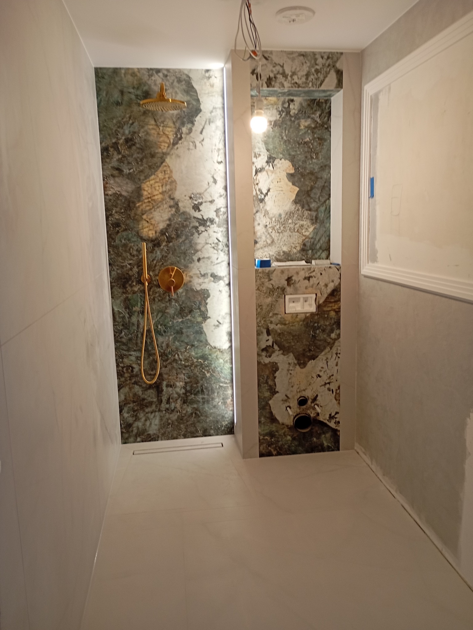 Wykończona łazienka z marmurową ścianą prysznicową z złotą armaturą i podtynkową toaletą. Widoczne białe płytki podłogowe i fragment instalacji elektrycznej.